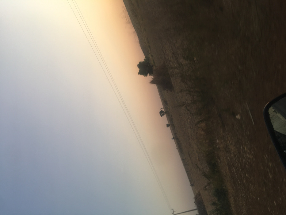 An angled view depicts a rural landscape under a hazy, gradient sky transitioning from blue to orange. A sparsely vegetated field stretches across the midground, dotted with a few trees. Power lines cut diagonally across the sky. The edge of a car, including a side mirror, is visible in the lower right corner, suggesting the image was taken from inside a vehicle. The atmosphere appears dusty or foggy.