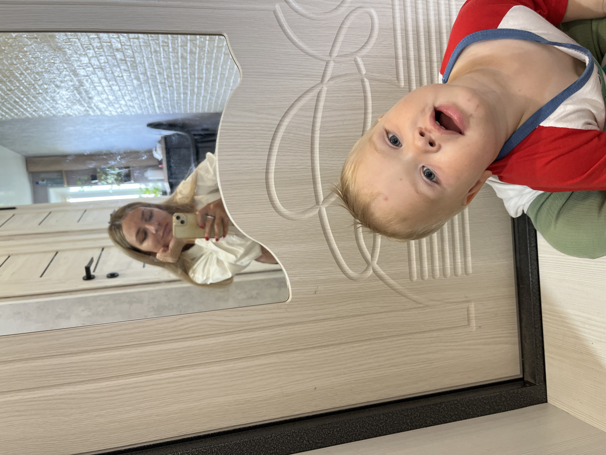 This media file shows a mirror reflection of a woman and a baby. The woman is holding a smartphone and taking a selfie. She has blonde hair and is wearing a white shirt. The baby has blonde hair and blue eyes, and is wearing a red and white shirt with blue trim. The background appears to be an interior with wooden doors and a textured wall. The image is oriented vertically, and the reflection suggests the subjects are in front of a mirrored surface, possibly a closet door. The overall mood is casual and appears to be a candid moment captured. The context mentions Chaikovsky, Russia, suggesting this is the location. There is no visible text in the image.