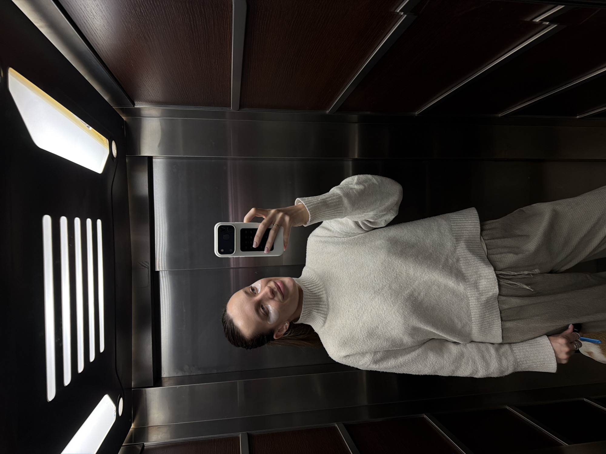A woman with eye patches is taking a selfie in an elevator. The elevator has a metallic panel with vertical light strips on one side and wooden panels on the top and bottom. The woman is wearing a light-colored turtleneck sweater and pants. The location is not specified in the image itself; Budva, Montenegro is only mentioned in the prompt.
