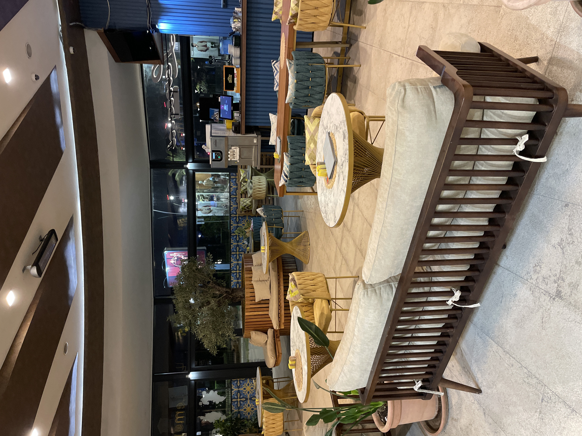 This image captures the interior of a modern and stylish cafe or restaurant in Budva, Montenegro, likely during the evening due to the illuminated indoor setting and darker views outside. The space is arranged with various seating options designed for comfort and aesthetics.
In the foreground and to the right, a long wooden sofa with a slatted back and light-colored cushions extends diagonally across the frame, providing ample seating. Its design is sleek, and white ties are visible securing the cushions. Adjacent to this, several round tables with white marble-patterned tops and intricately designed golden bases are paired with chairs featuring woven yellow or blue upholstery and matching cushions. Further back, a long, high wooden table with stools also upholstered in yellow and blue creates a bar-height dining or lounge area.
The bar section against the back wall is characterized by a distinctive blue corrugated surface. It features multiple screens, one of which is a black monitor displaying unreadable white text, possibly a menu or order interface. A white machine with "Carpe Diem" visible on its side, likely an ice cream or yogurt dispenser, is also present on the counter. Potted plants, including a small olive tree, are strategically placed to add natural elements to the decor.
The cafe is well-lit with various ceiling lights and wall sconces, creating a warm and inviting ambiance. Large windows span the length of the wall, looking out onto what appears to be a street or exterior area with reflections and distant lights. A neon sign with stylized, unreadable text is visible on the wall behind the bar area. The floor is tiled with light-colored, possibly beige or gray, square tiles. The cafe appears to be unoccupied, suggesting it might be either closed, awaiting customers, or captured during a quiet moment.