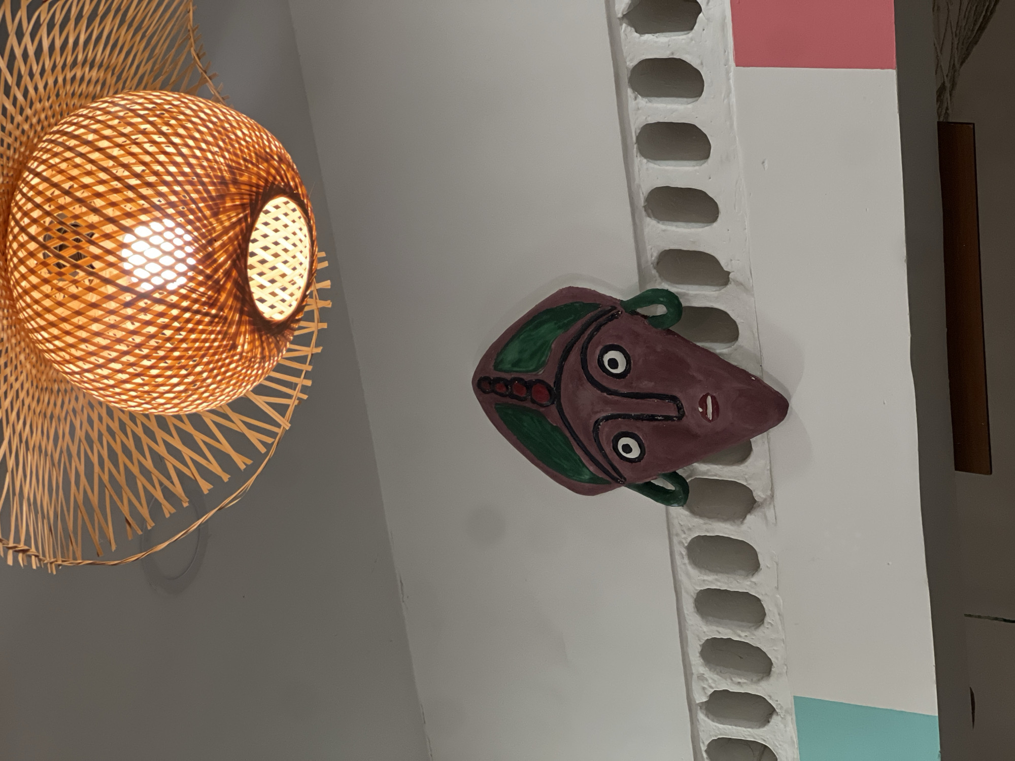 The image shows an interior space in Nha Trang Ward, Vietnam. A bamboo pendant lamp is on the left, emitting a warm glow. To the right of the lamp is a decorative mask mounted on a wall. The wall is white, with a textured, vertical column featuring oval indentations. The top of the column has a pink section, and the bottom has a light blue section. A dark brown vertical object is visible on the right side of the image, near the edge.