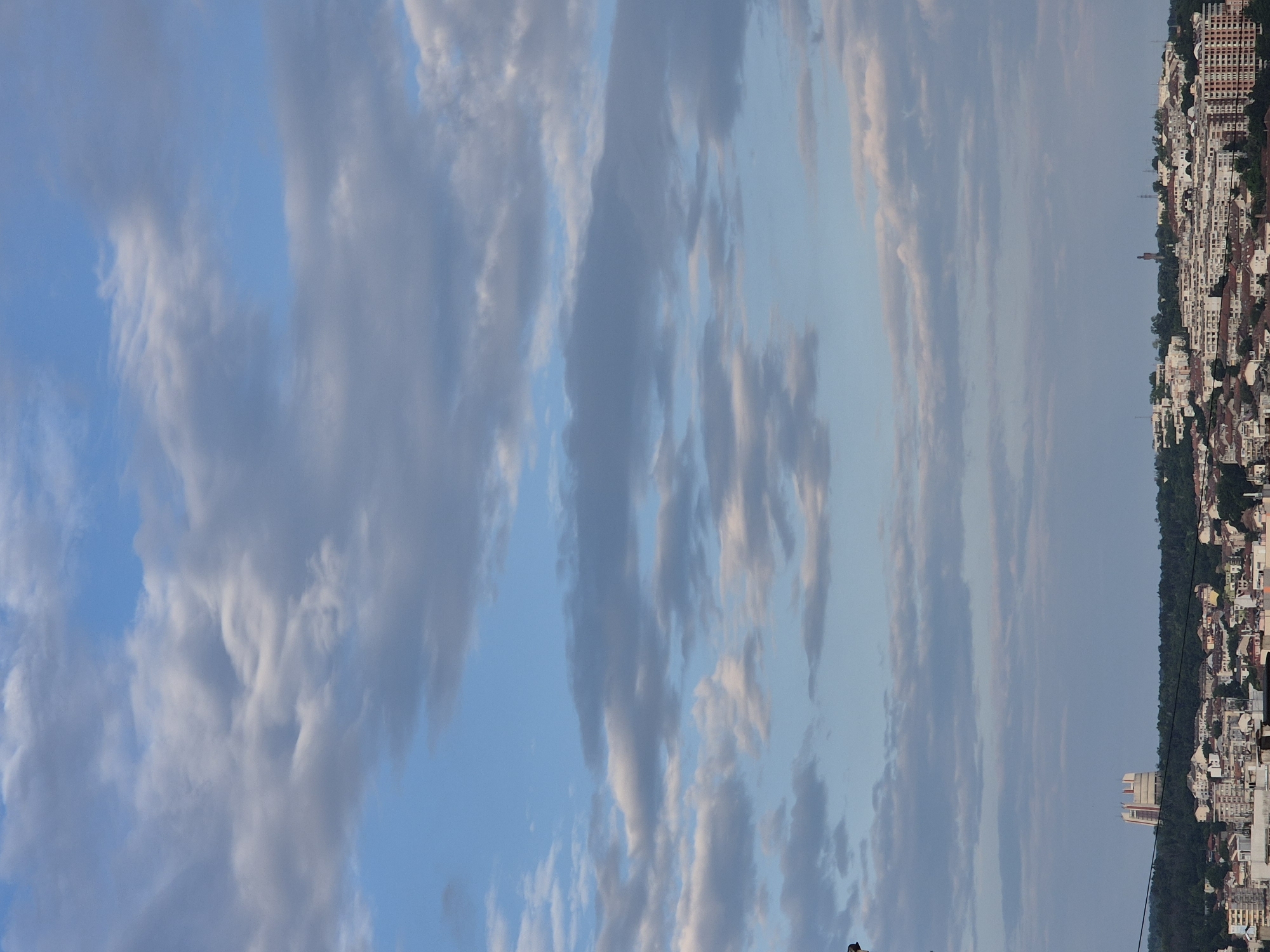This vertical shot captures an urban landscape under a cloudy sky. The upper two-thirds of the image are dominated by a pale blue sky interspersed with numerous stratocumulus and altocumulus clouds, exhibiting varied shades of white and grey due to differential sunlight and shadow. The clouds appear layered and somewhat turbulent. The lower third of the image depicts a sprawling city, situated on rolling hills densely covered with green trees and foliage. A prominent, light-colored, multi-story structure with a distinctive architectural design, possibly a water tower, stands on a hilltop towards the left. Residential buildings, varying in height and design, with terracotta, white, and grey roofs, cover the urban area. A dark, thin linear element, consistent with an overhead cable or power line, horizontally traverses the image just above the city's tree-covered skyline. No individuals or discernible activities are visible. The location is the city of Unknown, Unknown.