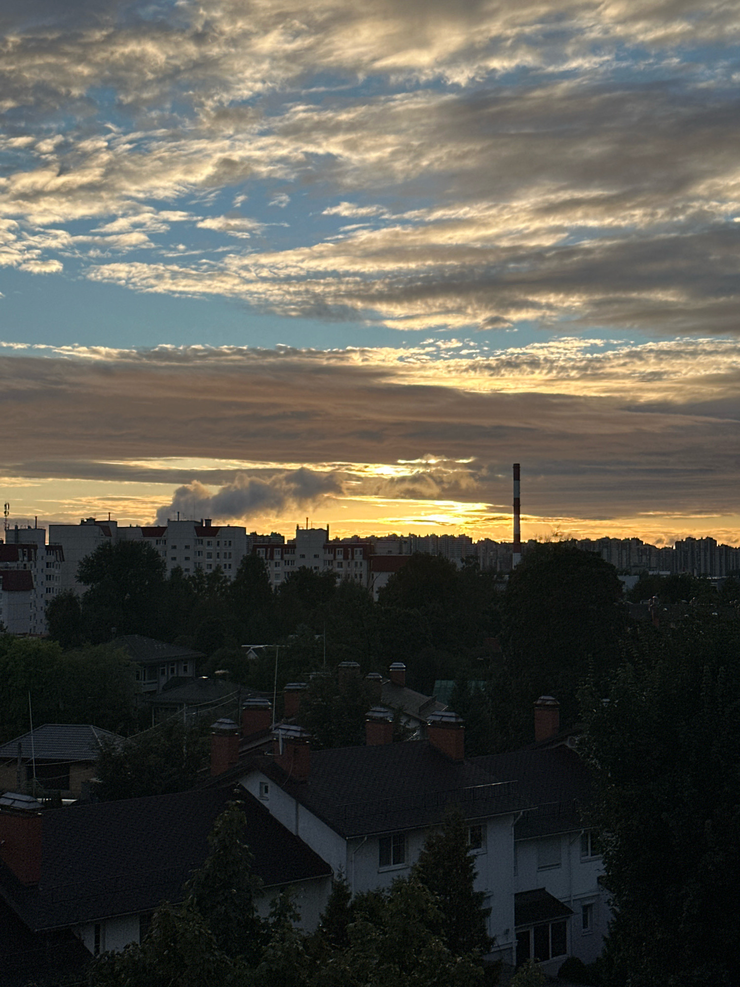 The image captures a dusky urban landscape with a striking sky. The sky is filled with layered, wispy clouds, predominantly in hues of grey, white, and a soft golden-orange near the horizon. The sun is partially visible, casting a warm glow through a break in the clouds. Below the sky, a city skyline is visible, characterized by rows of apartment buildings with red-tiled roofs. A prominent red and white striped smokestack stands tall among the buildings. In the foreground, the dark silhouettes of trees frame the scene, obscuring the lower portions of residential houses with dark roofs and visible chimneys. The overall impression is a city at the cusp of twilight, with the natural beauty of the sky contrasting with the man-made structures.