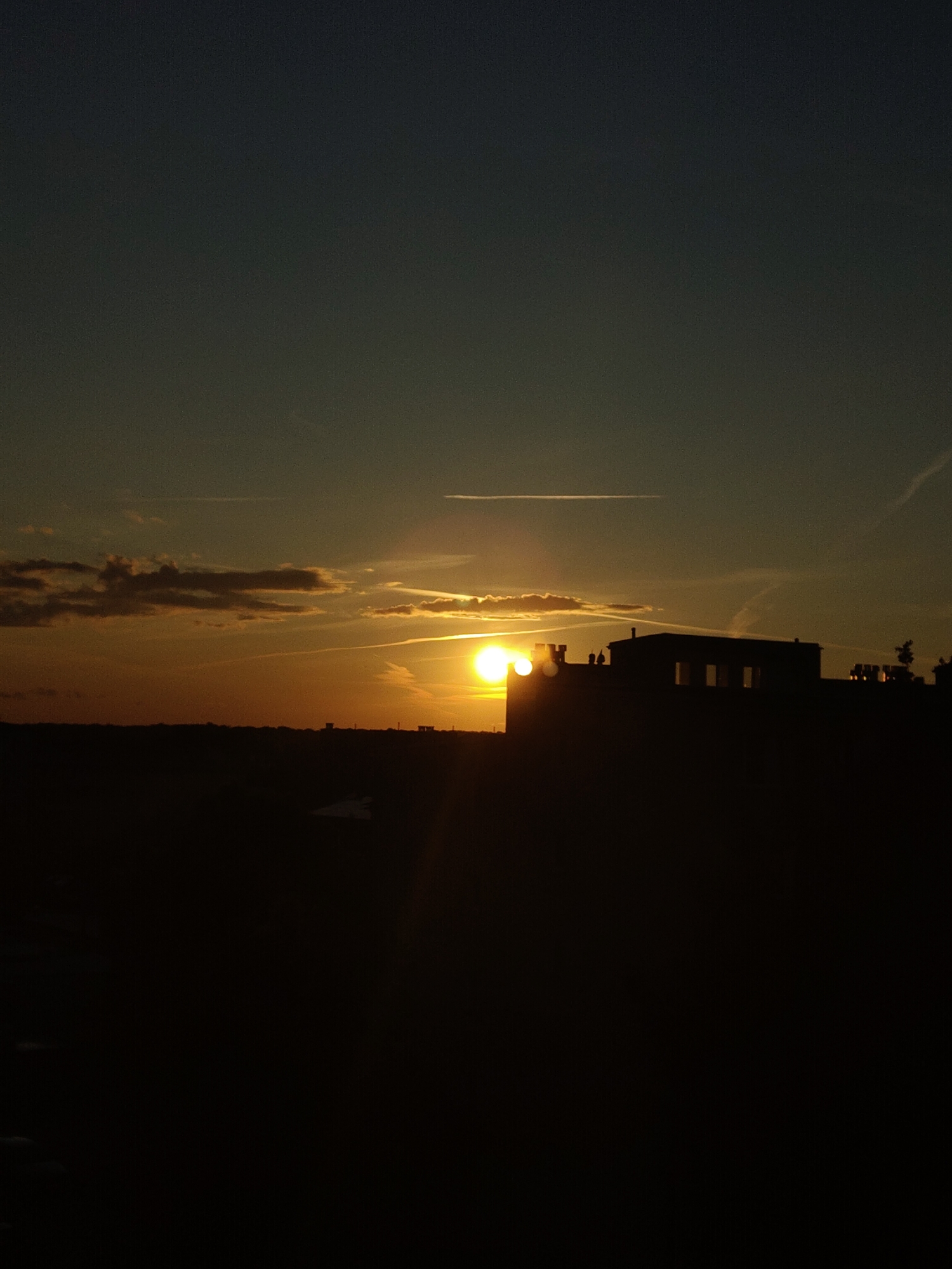 The image captures a silhouetted cityscape at sunset in Nowy Dwór Mazowiecki, Poland. The sun, a bright orb of light, is low on the horizon, casting a warm golden glow across the sky and illuminating wispy clouds. In the foreground, a dark silhouette of buildings dominates the lower half of the frame. These structures appear to be modern, with visible windows and rooftop features resembling crenellations. The surrounding landscape is also rendered in silhouette, creating a stark contrast against the illuminated sky. A faint, lighter patch on the ground in the lower center might indicate a reflective surface or a lighter colored object against the dark terrain. The overall impression is one of a quiet evening as the day transitions to night.