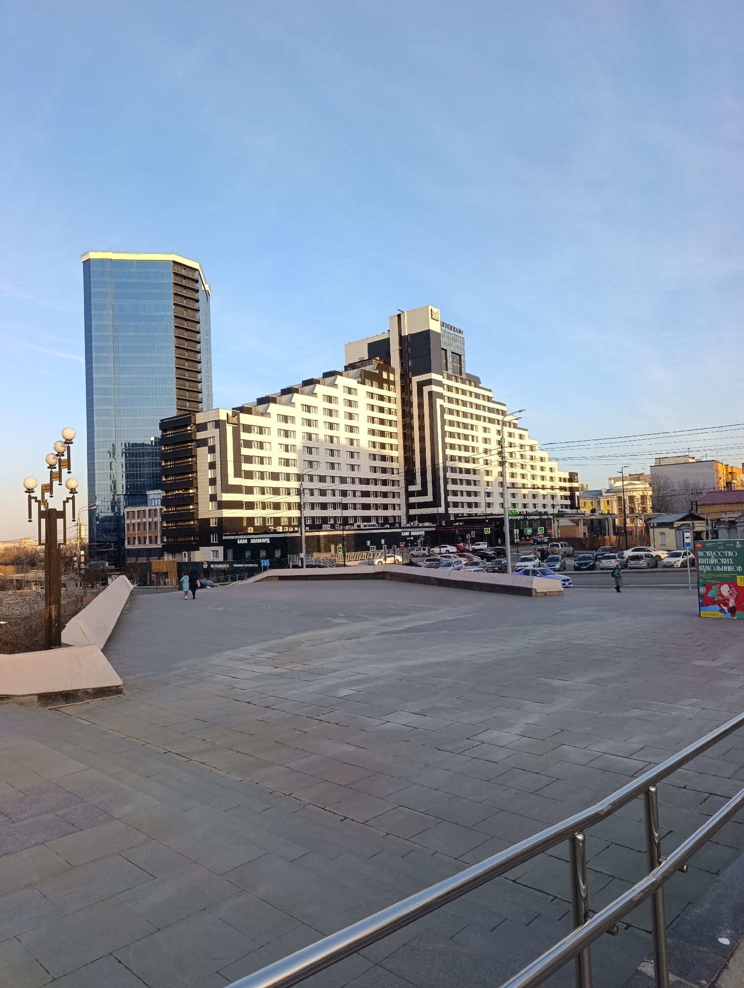 This image presents an urban landscape in Krasnoyarsk, Russia, featuring a combination of modern architecture and street activity under a clear, bright blue sky, indicative of a sunny day. The lighting suggests either morning or late afternoon, with sunlight casting clear shadows and illuminating the buildings from one side.
In the foreground, a spacious paved plaza, constructed from grey rectangular tiles, stretches out, bordered by a low concrete wall on the left and stainless steel railings on the right. Decorative streetlights with multiple spherical lamps line the left side of the plaza.
The scene is dominated by two distinct buildings. On the left, a tall, sleek skyscraper boasts blue reflective glass facades and dark, vertical architectural elements. To its right, a prominent stepped building with a unique design features white and black horizontal and diagonal patterns across its many levels, tapering upwards.
On the ground floor of the stepped building, several signs are visible, including "БАНК АВАНГАРД" (Bank Avangard), "SLOGAN," and "ПРОДАЖИ КВАРТИР" (Apartment Sales). The word "PREMIUM" and another logo are also visible near the top of this building. Reflected on the blue skyscraper, "ПРОДАЖИ КВАРТИР" can also be discerned.
The middle ground shows a busy street with numerous parked and moving cars. Several pedestrians are visible, including two individuals walking on the paved plaza and others on the street and sidewalks. On the far right, a green advertising board displays the text "ИСКУССТВО КИТАЙСКИХ КУКОЛЬНИКОВ" (The Art of Chinese Puppeteers) with a partial date "3.1-". A bus stop sign with "Остановка" is also present. The scene depicts a lively urban environment with a mix of contemporary and functional elements.