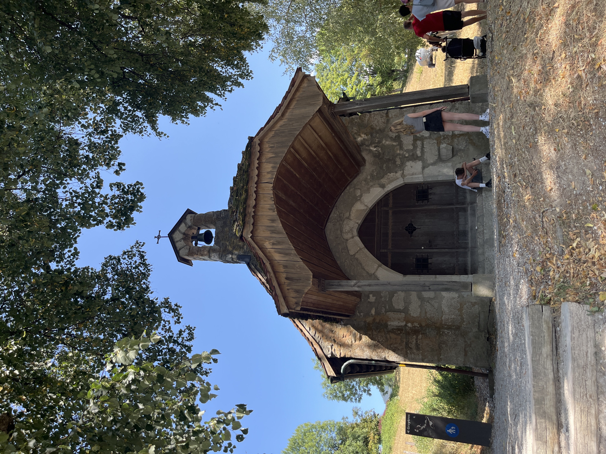 The image shows a small stone chapel with a wooden roof and a bell tower. The chapel is situated on a grassy slope under a clear blue sky, with trees providing shade. In front of the chapel, there are several people. A young person is sitting on the steps, appearing tired. Two young women are standing near the entrance, one looking away from the camera. Further to the right, a man in a red shirt and another person in a white shirt are visible, with a stroller nearby. A signpost indicating "Gruyères" is located to the left of the chapel. The ground in front of the chapel is covered with gravel and dry leaves. The overall setting suggests a rural or semi-rural location in Pringy, Switzerland.