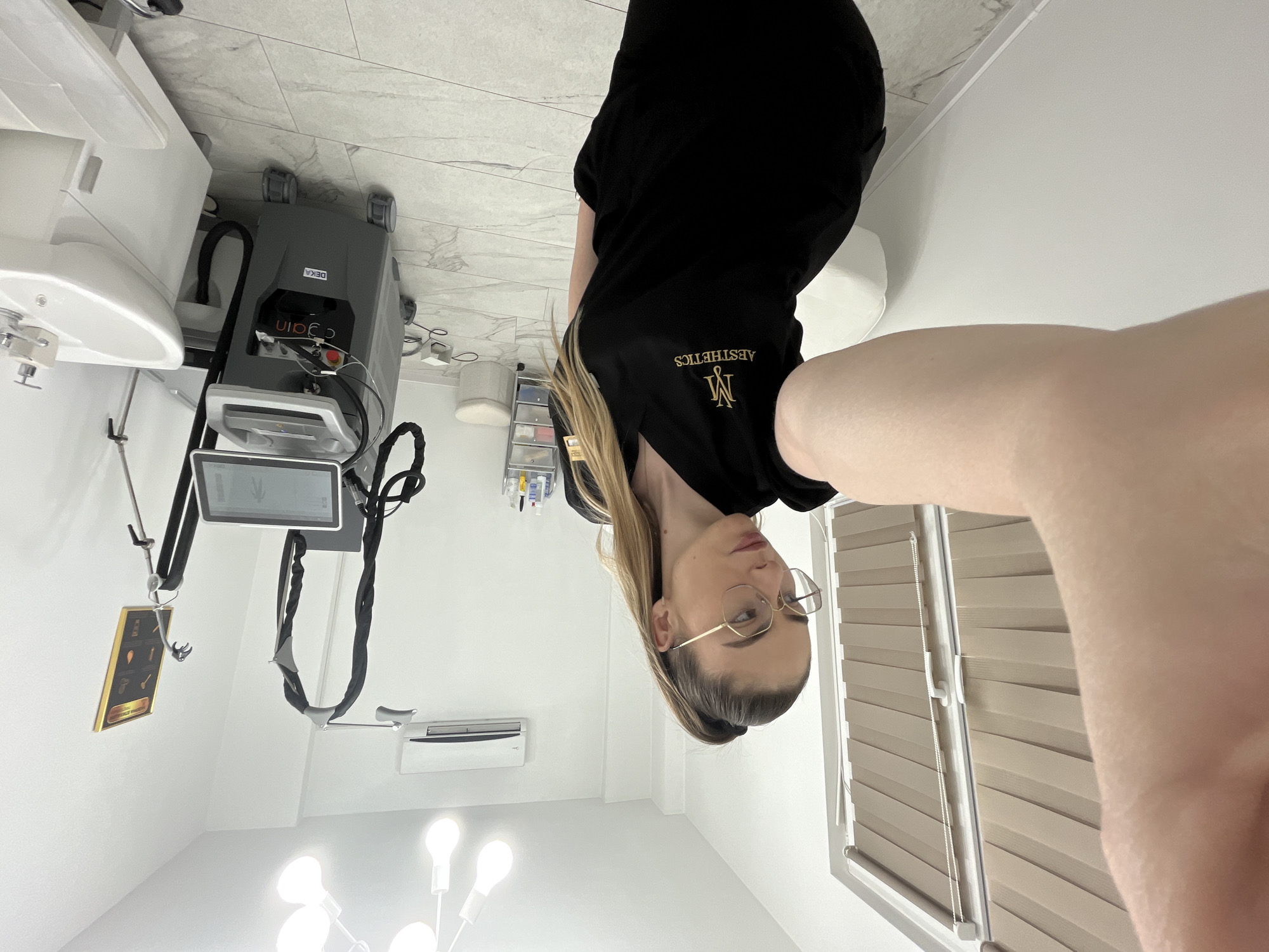 The image depicts a woman taking a selfie in what appears to be a medical aesthetics clinic. The woman has light hair, is wearing glasses, and is dressed in a black uniform with the word "Aesthetics" printed on it.
In the background, there is medical equipment including a DEKA laser machine, white medical equipment, a wall-mounted air conditioner, a light fixture, blinds and a poster that contains medical information. The location is unknown.