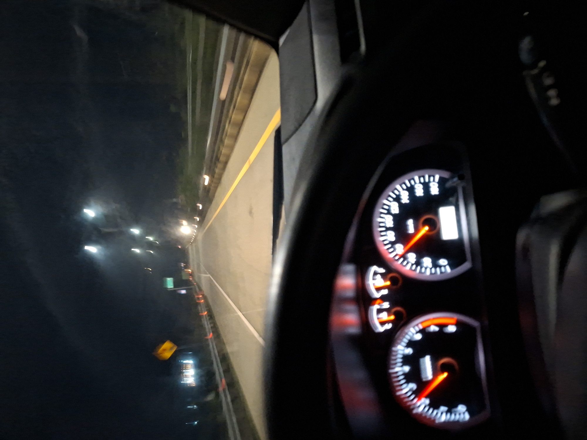 The image captures a perspective from inside a vehicle driving at night, with the view divided between the car's interior and the external road.
On the right side, the car's dashboard and a portion of the steering wheel are visible. The instrument cluster is brightly illuminated, featuring at least two primary circular gauges with black faces, white markings, and prominent red needles. The upper gauge appears to be a tachometer, indicating RPMs with numbers ranging from 1 to 7. The lower gauge is likely a speedometer, showing speed in increments from 20 to 220. Below the tachometer, there are smaller, red-lit indicators, possibly for fuel and temperature levels.
The left side of the image shows the dark night environment outside the car. The sky is black, and the road ahead is illuminated by a series of streetlights and other vehicle lights, which appear as blurred, elongated streaks of white and yellow due to motion or long exposure. A clear yellow line marks the edge or center of the road, running parallel to the vehicle's path, along with some white road markings. On the roadside, there are several orange reflective cones or barriers, and two indistinct road signs—a green rectangular one and a yellow diamond-shaped one—are barely visible, with no legible text.
The scene depicts nighttime travel on a well-lit road or highway, conveying a sense of ongoing movement in Aron Bunoet, Indonesia.