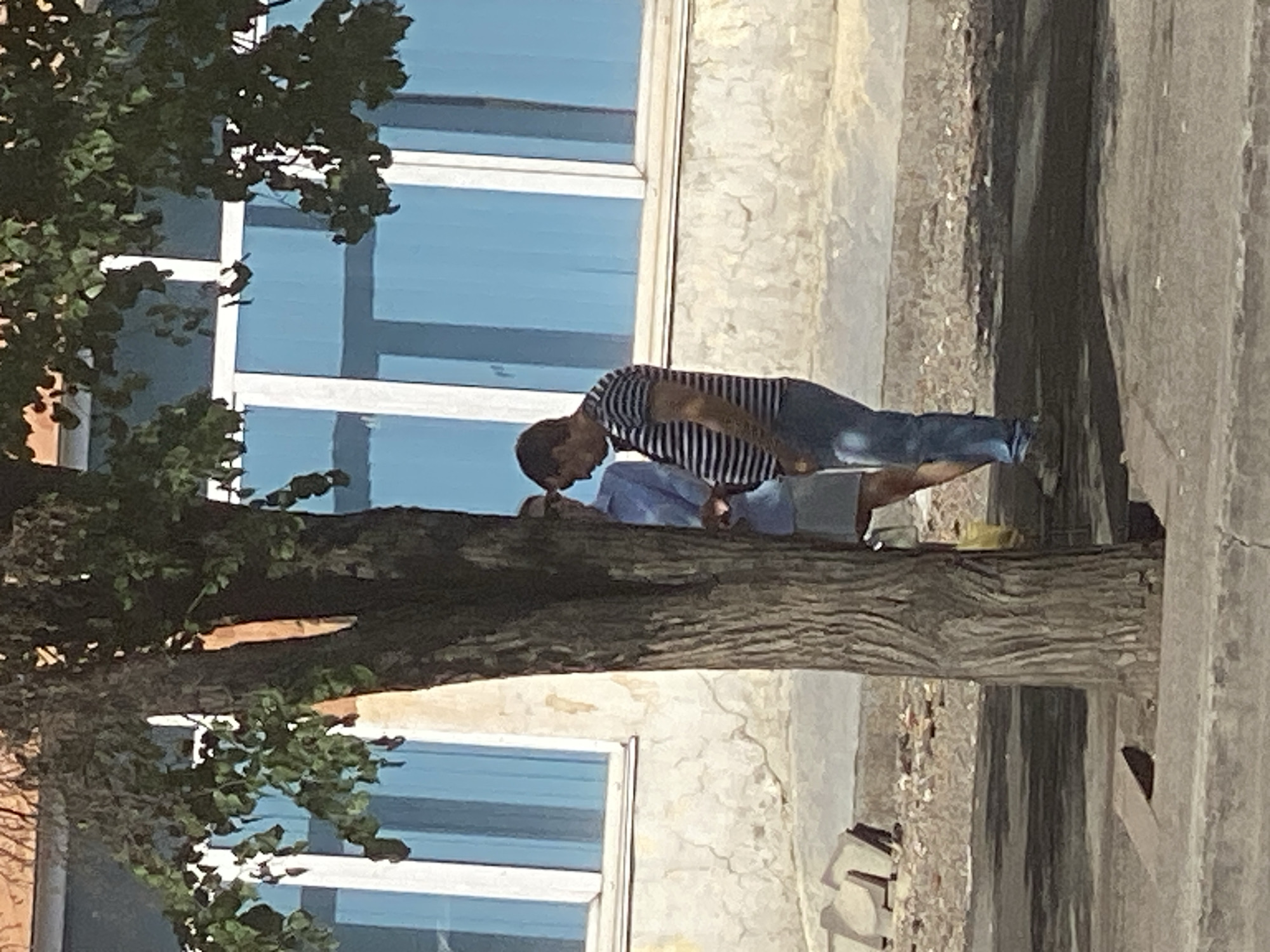 Two individuals are standing next to a tree in front of a building with blue-tinted windows. The person on the right, a male, is wearing a striped t-shirt and jeans and appears to be looking down. The person on the left, partially obscured by the tree and the male, is wearing a light blue shirt and a light-colored skirt. The ground in front of the building is paved, with debris scattered around. The building has a light-colored, textured facade. The overall scene suggests an outdoor urban environment. Without further information or context, it is not possible to determine the specific location within Novokuznetsk, Russia.