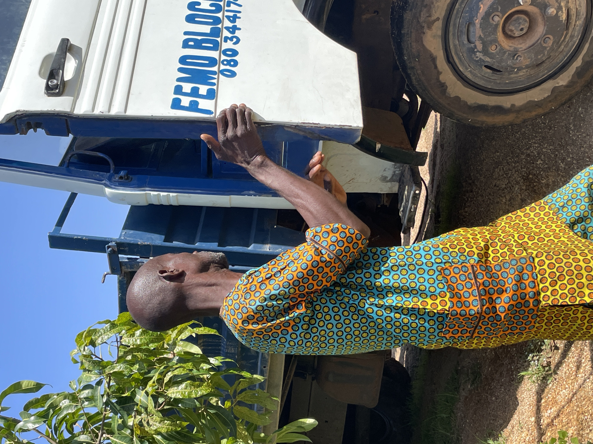 A man wearing a vibrantly patterned shirt and trousers, featuring a bright yellow base with circular motifs in orange, teal, and black, stands with his back to the camera, facing a white truck with blue trim. His right hand is on the open door of the truck, and his left arm is bent, with his hand resting on his side. The truck has blue lettering on its side that reads "FEMO BLOCK" followed by a phone number, "080 34417". The scene appears to be outdoors, with a clear blue sky overhead and lush green foliage on the left side of the frame, suggesting a location with vegetation. The ground is covered in gravel and dirt. The overall setting suggests a commercial or logistical context, possibly related to construction or delivery services. The man's attire and the presence of the truck are the primary focal points of the image. The image is taken in Jos, Nigeria.