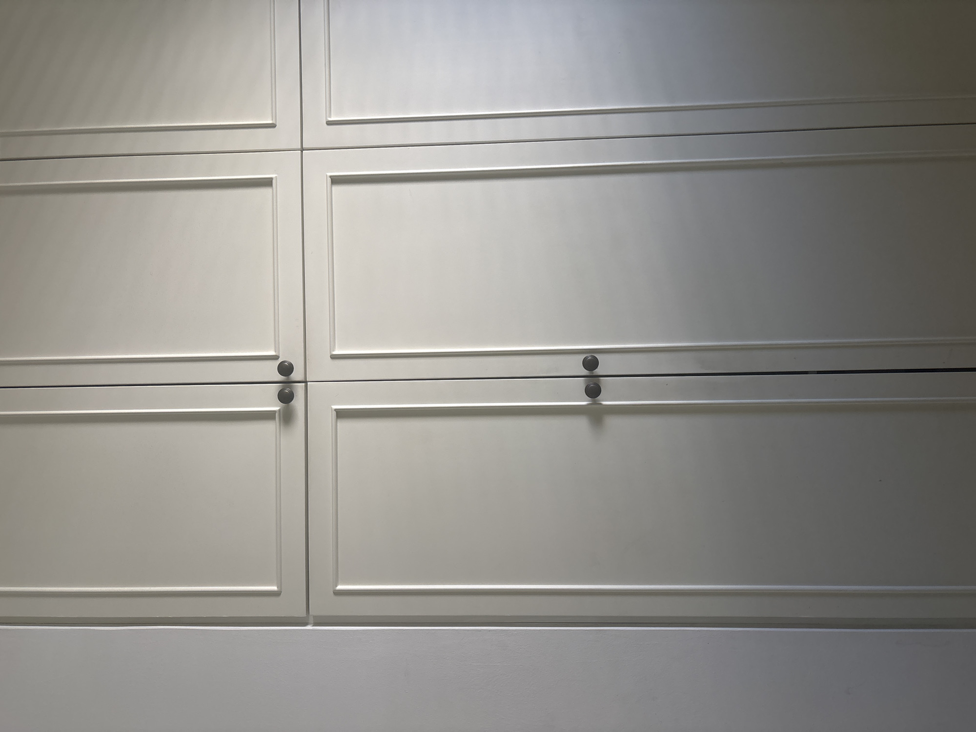 A close-up, eye-level, medium shot captures a section of a white closet with double doors on the top and bottom. The closet doors have a paneled design with dark, circular knobs for opening. The lighting creates subtle shadows, highlighting the texture of the closet doors and a wall to the left, which appears to be a lighter shade of white. The image does not show any people or actions. The location is identified as Lod, Israel.