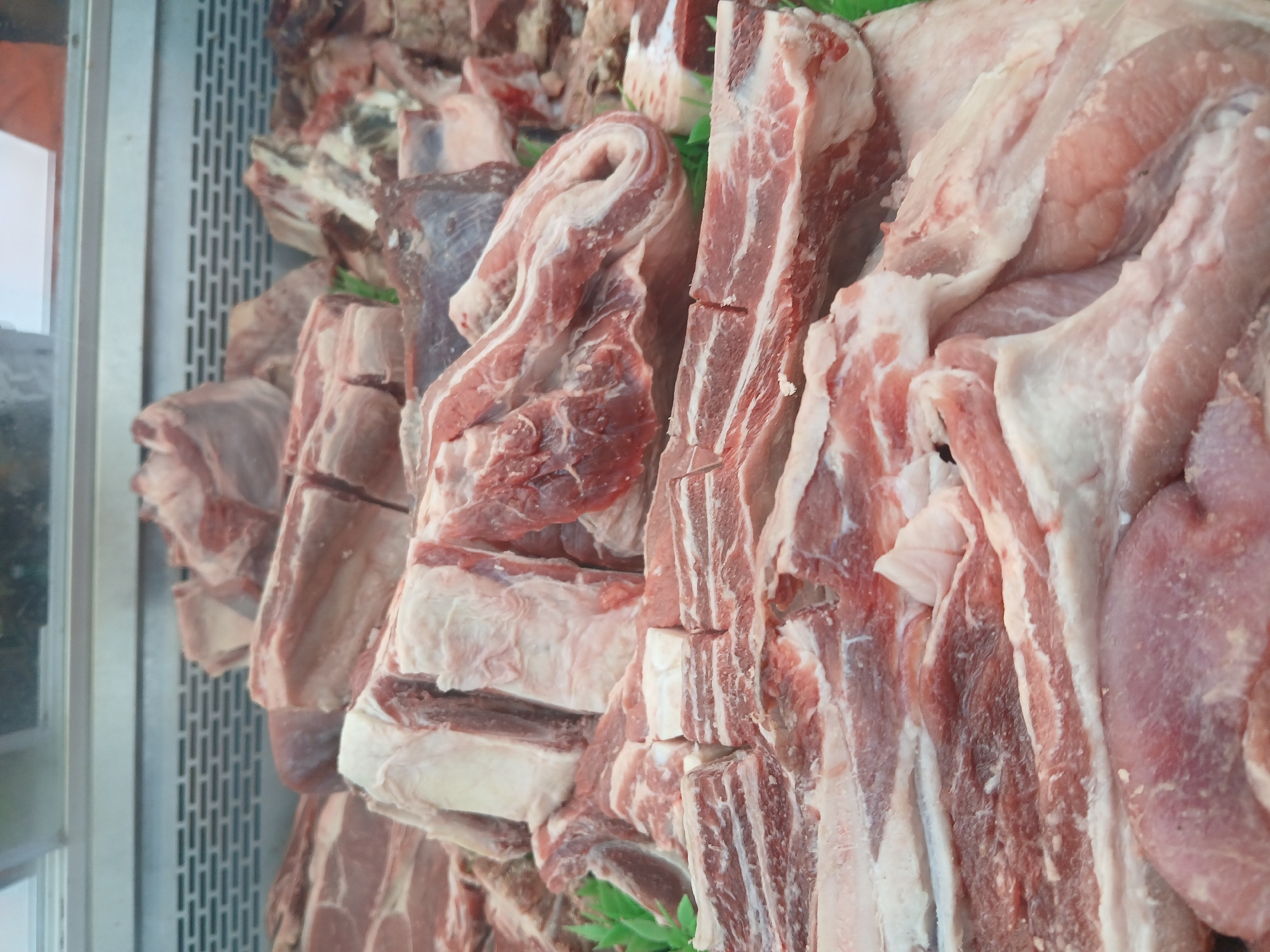 The image depicts a display of raw meat cuts, likely in a butcher shop or supermarket in Ciudad Guayana, Venezuela. The cuts include various types of beef and pork ribs. The meat is predominantly red and pink with visible fat marbling. The presentation is unorganized, with cuts piled on top of each other. A metallic rack is visible on the side, indicating the meat is displayed inside a refrigerated case.