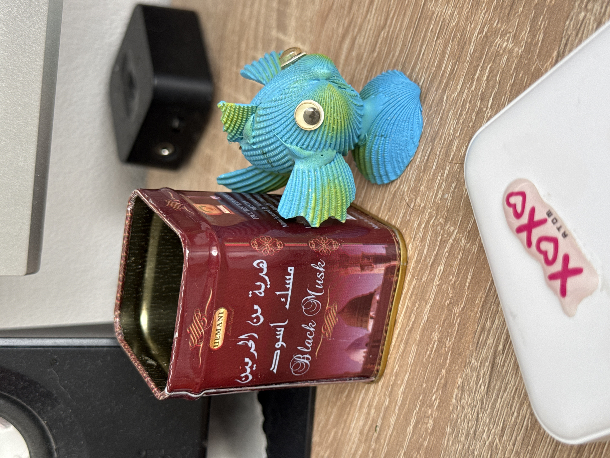 A fish-shaped figurine made of shells sits atop an open, empty maroon-colored tin labeled "HEMANI Black Musk" with Arabic script. To the left of the tin is a black, rectangular device. To the right of the tin is a white, rectangular object with a "XOXO" sticker on it. The objects are on a wooden surface.