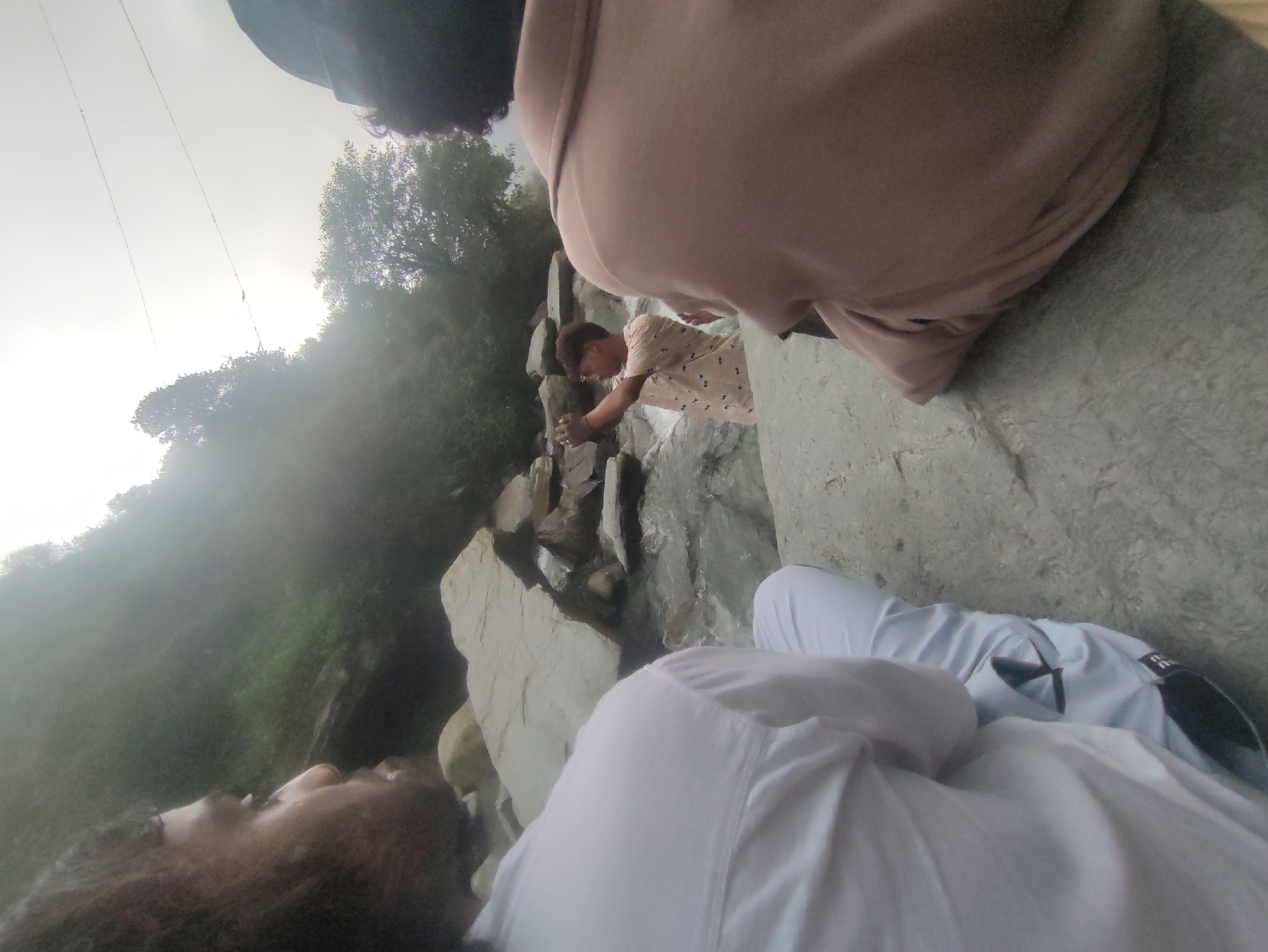 In the city of Bhagsu, India, three individuals are present near a rocky stream. On the left, a person wearing a white shirt and light-colored pants is partially visible, with their back to the camera, facing a misty, forested hillside. In the center, a young person in a patterned shirt is actively taking a photograph with their phone, facing the stream. To the right, another individual is seated with their back to the camera, wearing a brown jacket and a dark hat. The environment is characterized by large rocks, flowing water, and dense vegetation, with a hazy, overcast sky.