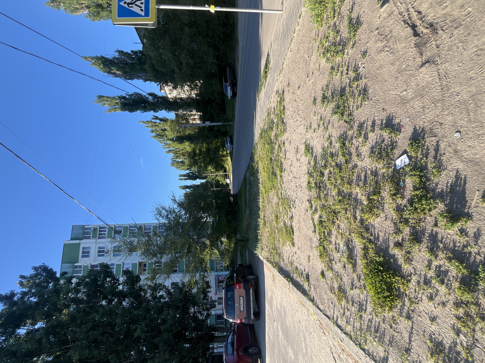 The image depicts a street scene in Voronezh, Russia, on a clear, sunny day. In the foreground, a patch of sandy ground with sparse green vegetation and some litter is visible, bordered by a concrete sidewalk. A dark-colored car and a red car are parked on the left side of the frame.
Behind the parked cars, a multi-story apartment building with a white and green facade stands tall. Several trees with dark green foliage line the street, some appearing tall and slender. Further down the road, other cars are parked or driving. A pedestrian crossing sign is mounted on a pole on the right side of the image. The sky is a vibrant blue, with power lines stretching across it. The overall impression is one of an ordinary urban environment in good weather.