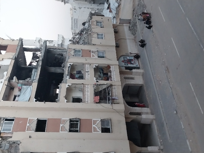 The image captures a seven-story building in Khan Yunis, Palestinian Territory, exhibiting severe structural damage in its mid-upper sections. The third, fourth, and fifth visible floors from the ground up are heavily damaged, with large portions of exterior walls and interior structures missing, exposing debris and rebar. The two lowest floors and the uppermost visible floor show less extensive damage, retaining balconies, some with clothes hanging, and some intact windows. A satellite dish is visible on an upper balcony.
To the right of the main building, another structure is severely damaged, with large piles of rubble in front of it. A white minaret-like structure, likely part of a mosque, is visible beyond the damaged buildings in the background.
In the foreground, a paved street with two white dashed lane markings runs horizontally, bordered by a concrete sidewalk. Debris is scattered across both the street and sidewalk. On the ground floor of the main building, arched entryways are present, and a commercial sign featuring a "SQUID GAME" logo is visible on the right side.
On the sidewalk near the commercial sign, a group of approximately eight individuals, predominantly men and youths, are gathered, some squatting or seated. Two individuals are partially visible within an arched entrance on the ground floor, one wearing a red garment. The sky above is overcast.