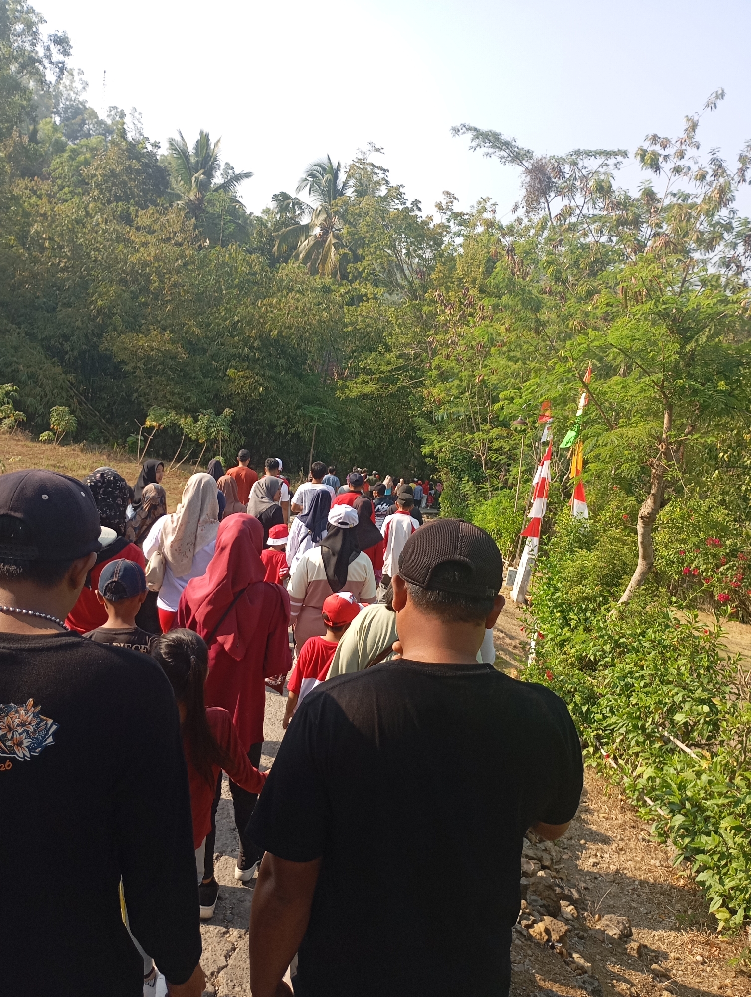 A group of people are walking along a dirt path lined with trees and vegetation. The path appears to be in a rural or semi-rural area. The sky is clear and bright. Several people in the group are wearing red and white clothing, and some are wearing headscarves. Small flags are visible on poles along the side of the path. The image was taken in Ketepung, Indonesia.