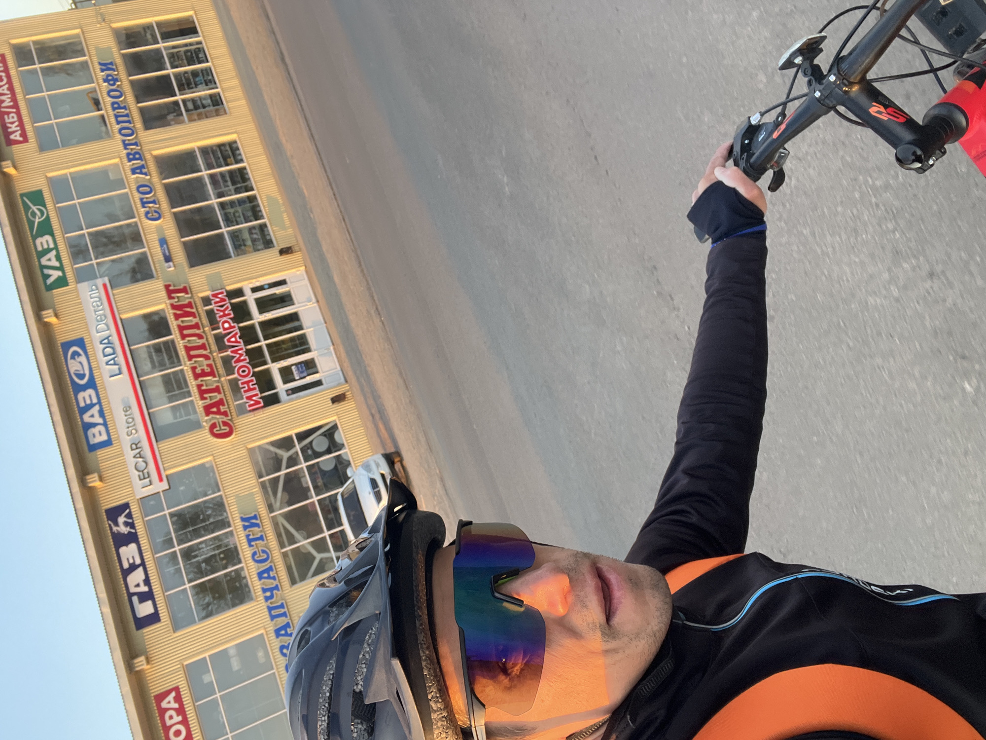 Here is a detailed description of the image:
**General Scene:**
The image appears to be a selfie taken by someone riding a bicycle on a paved road. The photograph is angled, capturing the rider, the handlebars of the bike, and the surrounding environment.
**People:**
A person is visible in the lower right of the frame. They are wearing a bicycle helmet and mirrored sunglasses. The person is also wearing a long-sleeved cycling jersey with black and orange colors.
**Subjects:**
* **Bicycle:** Part of the bicycle is visible, including the handlebars and a red water bottle.
* **Building:** There is a multi-story building in the background. It appears to be a commercial building, with several large windows and signage in Russian.
* **Road:** The road is paved and slightly slanted.
**Objects:**
* **Helmet:** A gray cycling helmet is on the person's head.
* **Sunglasses:** The person is wearing mirrored sunglasses.
* **Water Bottle:** A red water bottle is attached to the bicycle frame.
* **Signage:** The building has several signs with various business names and logos.
**Actions:**
The person is riding a bicycle and has taken a selfie.
**Interactions:**
The person is holding onto the bicycle handlebars. The building serves as a backdrop for the selfie.
**Environment:**
The scene appears to be an urban or suburban area with a commercial building. The lighting suggests it could be morning or late afternoon.
**Location (Chaikovsky, Russia):**
Based on the signage on the building, the location is likely in Russia. Specifically, the signage contains names of Russian automobile brands like VAZ (also known as Lada), GAZ (Gorky Automobile Plant), and UAZ (Ulyanovsk Automobile Plant). Also, there is a sign "САТЕЛЛИТ-ИНОМАРКИ", which translates to "Satellite - Foreign Cars", which refers to imported cars, making it likely a car part retail store.
Based on the presence of the signage, one can deduce the location is in a Russian city. Determining specifically Chaikovsky requires more information from the environment.
Overall, the image captures a moment during a bicycle ride in an urban setting, likely Russia.