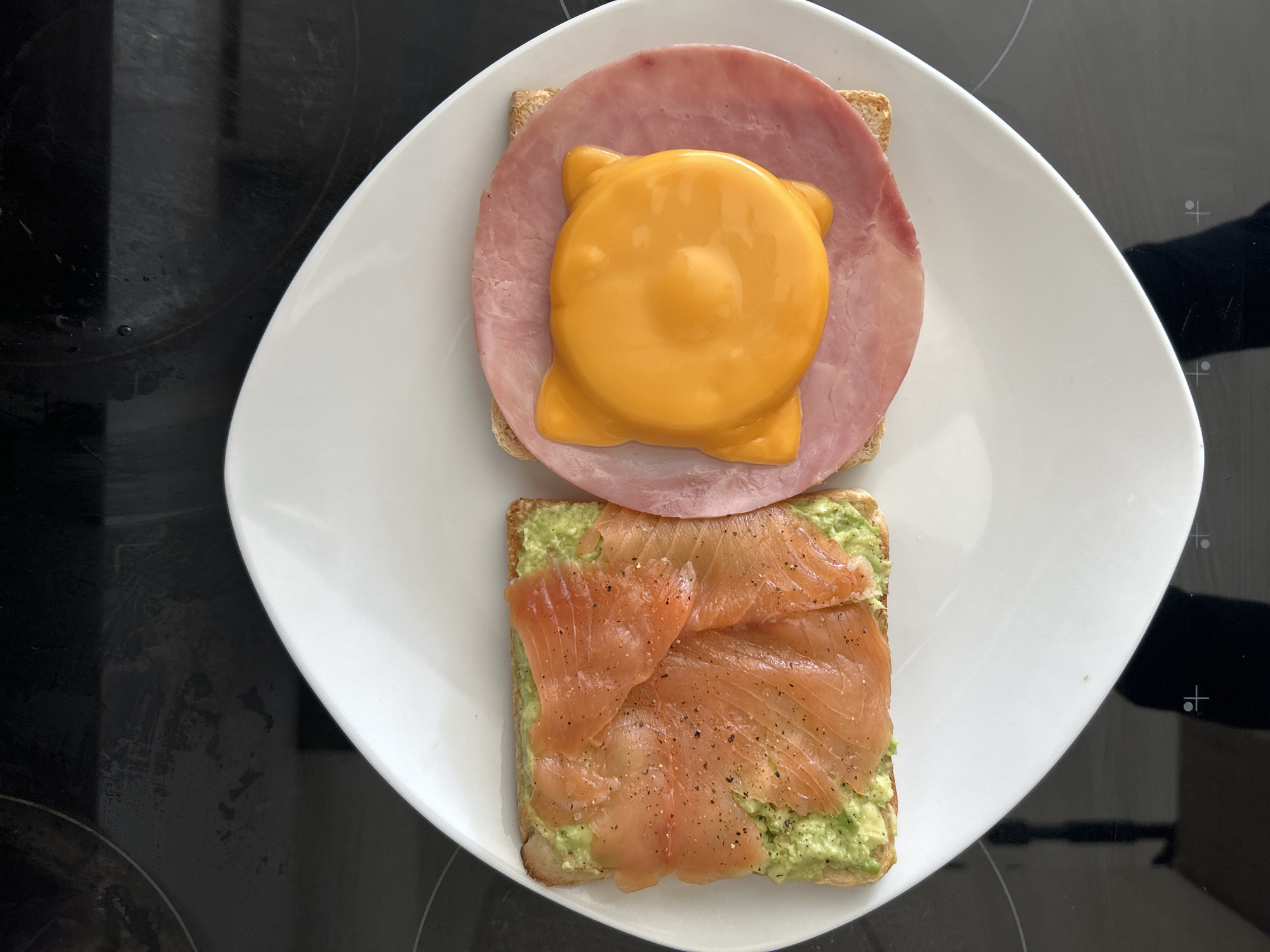 The scene is an overhead view of a meal prepared on a dark, reflective kitchen stovetop, indicating a domestic setting in Unknown, Unknown. No individuals are present. A white, oval-shaped plate occupies the center of the frame, holding two distinct portions of toast.
On the left side of the plate, one piece of rectangular, lightly toasted bread is topped with a layer of mashed avocado, which is then covered by several folded slices of smoked salmon. Black specks, likely ground pepper, are visible across the salmon and avocado.
On the right side of the plate, a second piece of rectangular, lightly toasted bread serves as a base for a large, circular slice of pink processed ham. Centered on the ham is a bright yellow, circular object with four distinct rounded protrusions and a raised central dome, consistent with a melted, molded slice of processed cheese.
The dark stovetop background shows subtle circular markings of heating elements and various smudges and reflections. The food appears freshly prepared and ready for consumption.