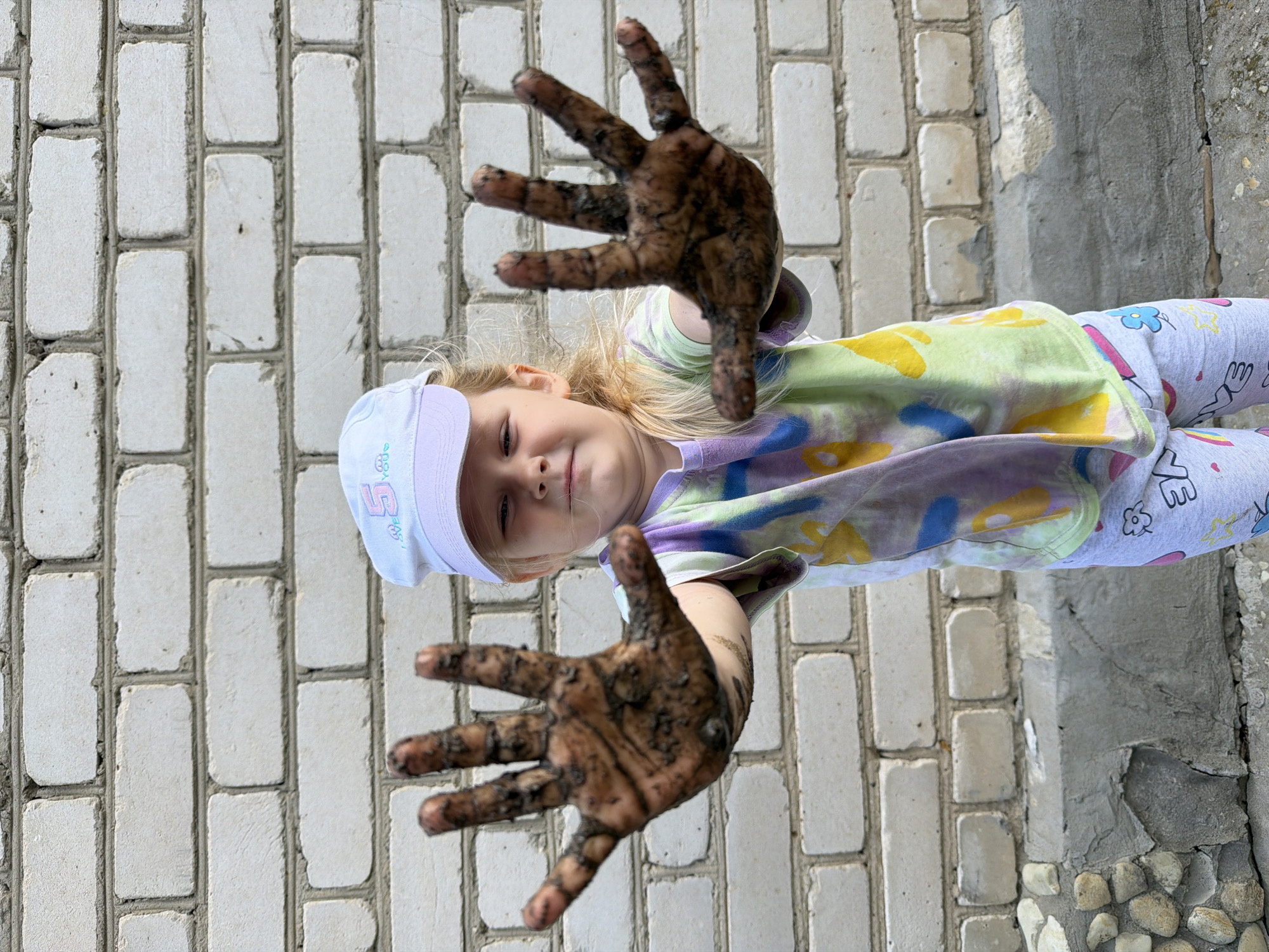 A young girl with blonde hair and wearing a white baseball cap, a tie-dye t-shirt, and light grey leggings with colorful drawings is standing in front of a white brick wall. Her hands, covered in mud, are extended towards the camera. The background consists of the brick wall and a small section of textured ground at the bottom. The image appears to be taken outdoors, possibly in a residential area of Kamyshin, Russia, judging by the style of the brickwork and general outdoor setting.