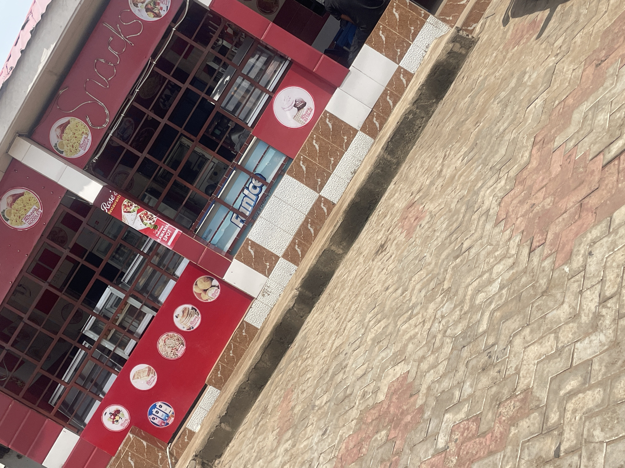 The image shows the exterior of a restaurant called "Rose's Snacks" located in Lokoja, Nigeria. The building has a red facade with metal grates covering the windows. Various circular stickers displaying food items such as noodles, pancakes, and ice cream are visible on the red surface. Above the windows, "Snacks" is written in neon lights. The ground in front of the building is paved with interlocking bricks. The scene appears to be during daylight.