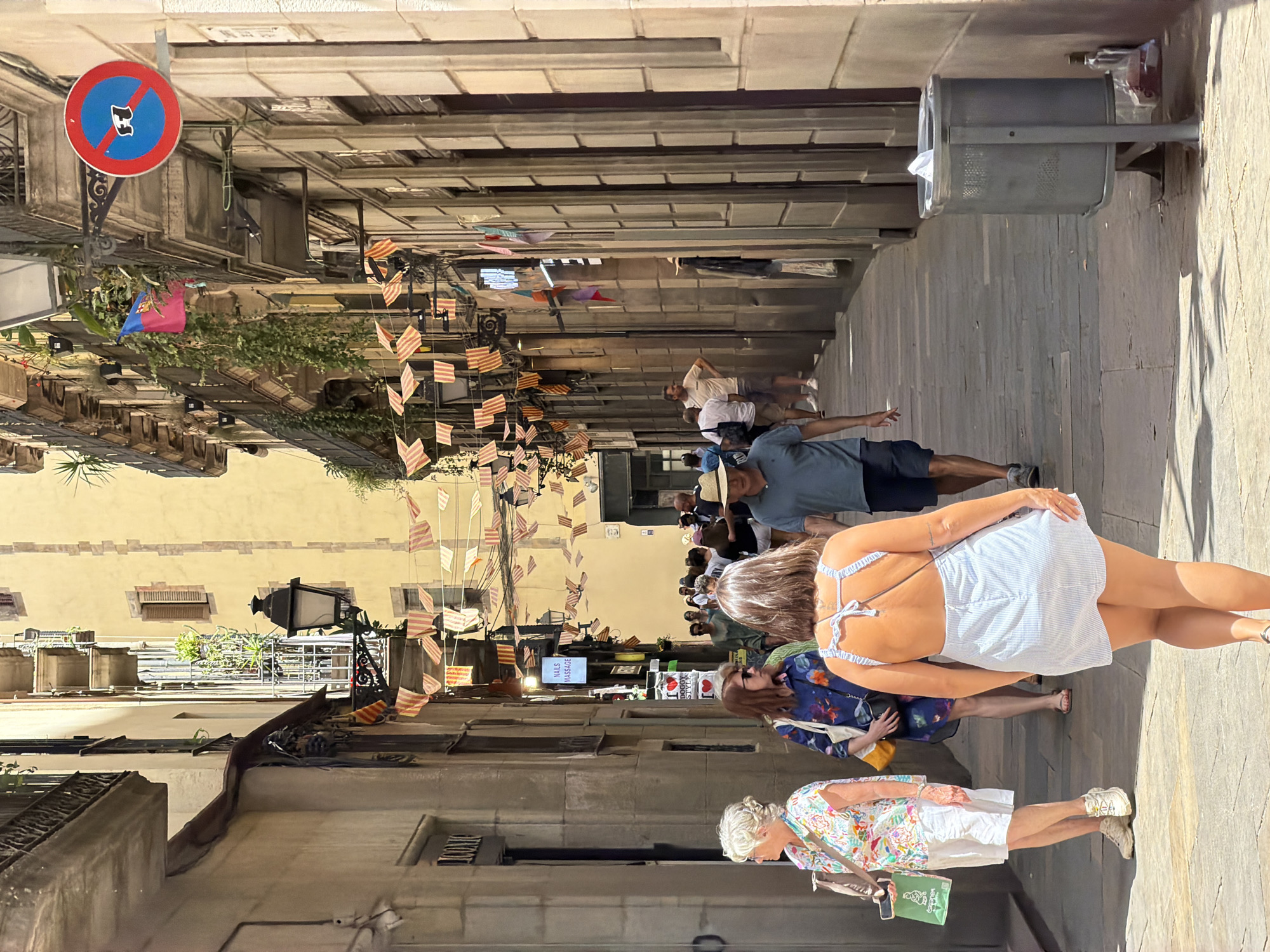 The image depicts a sun-drenched, narrow street in Barcelona, Spain, likely in the Gothic Quarter, as evidenced by the architecture. The street is lined with tall buildings featuring balconies and ornate details. String pennants, likely the Catalan flag colors of red and yellow, are draped across the street overhead.
In the foreground, walking away from the camera, is a woman wearing a light blue and white striped mini-dress with an open back. To her left, a woman with short, blonde hair wears a colorful floral print top and white shorts, holding a green shopping bag. To the left of that, another woman in a dark floral print dress with a yellow purse slung over her shoulder walks with a slight bend forward.
Further down the street, a man wearing a straw hat and a dark blue t-shirt walks towards the camera, his left hand outstretched. Behind him, a man in a grey t-shirt and dark shorts walks away, his left hand extended. Other pedestrians are visible further down the street, in various states of motion. Shops are visible along the sides of the street, with signage including "ICONIC" and "NAILS MASSAGE." A street lamp is mounted on the left side of the street, and a "no parking" sign with a symbol is visible on the right. A metal trash can is situated on the right side of the street near the bottom right corner of the image. The ground is paved with stone slabs, and shadows from the buildings are cast across the pavement.
