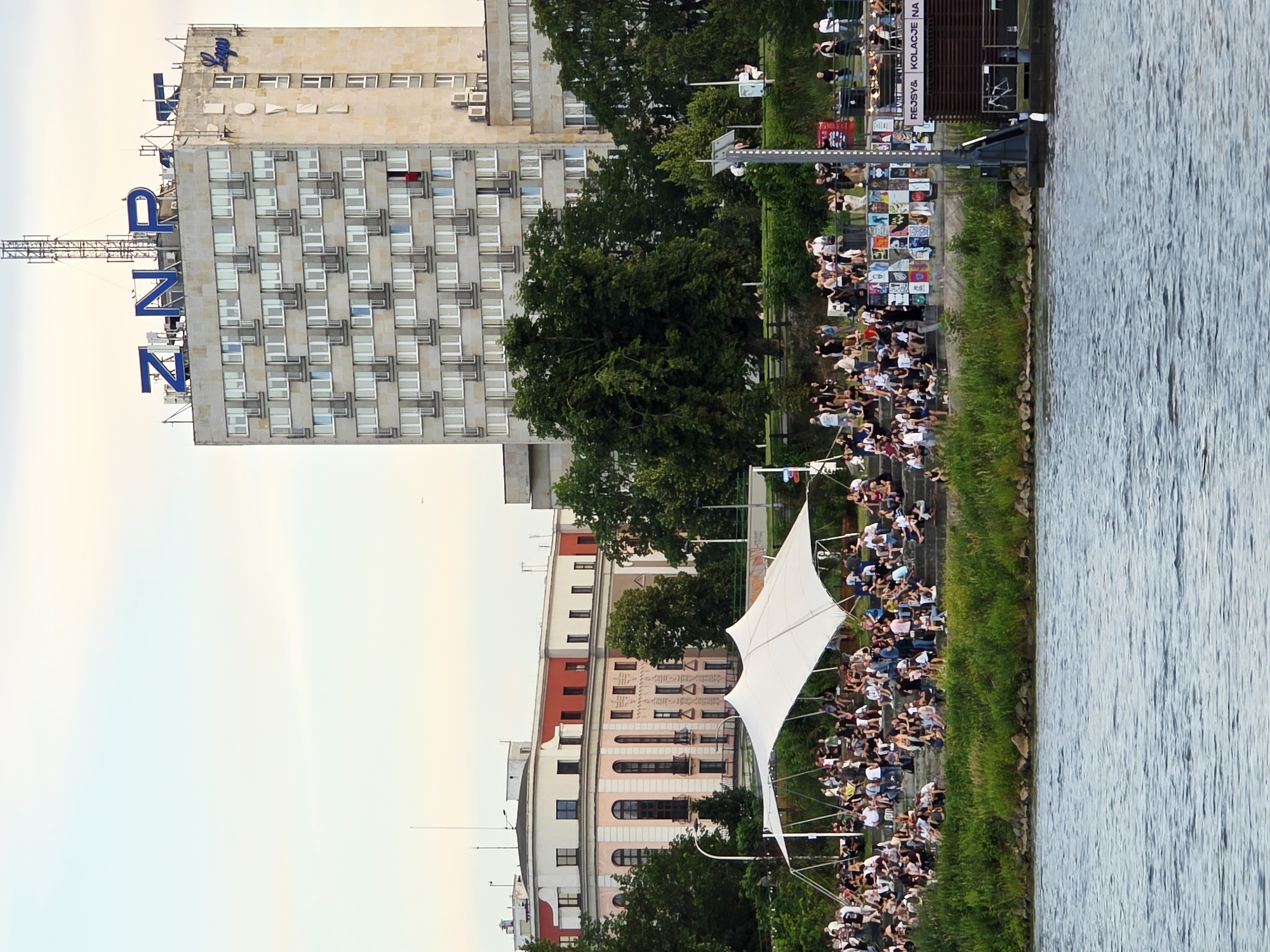 This is a full-shot, eye-level, outdoor photograph of a waterfront gathering in Warsaw, Poland. In the foreground, a wide, blue river occupies the bottom portion of the frame. The middle ground features a densely populated grassy embankment leading up to a tiered seating area made of concrete steps. A large, white, tensile canopy is erected over the seating area, providing shade for the numerous people gathered there, who are sitting and standing, facing towards the river. Behind the seating area, a cluster of trees stands, partially obscuring a pink and white building with arched windows. Dominating the background is a tall, grey, multi-story building with a sign that reads "ZNP" in large blue letters. The building also has a sign that appears to read "HOTEL" on its side. The sky above is overcast and pale.