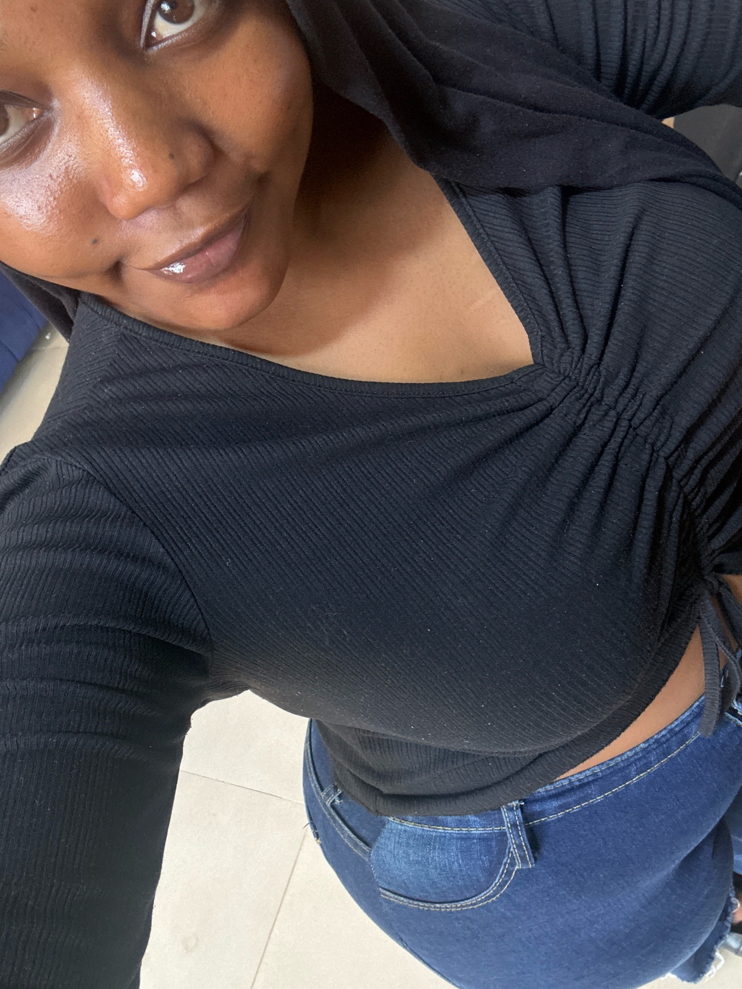 The image is a close-up, overhead shot of a Black woman wearing a black, ruched, long-sleeved top and blue jeans. The woman's face is partially visible in the upper left quadrant, with her eyes looking towards the camera. Her lips are glossed. The background appears to be an indoor setting with tiled flooring, possibly in Jos, Nigeria, though no specific landmarks are identifiable.