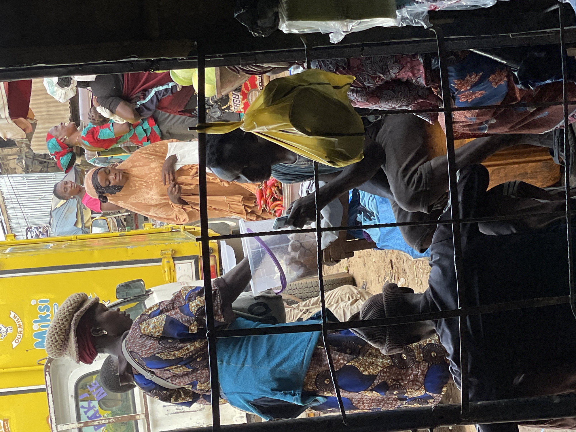 This media file captures a crowded scene inside a vehicle, likely a bus or truck, filled with people and their belongings. The orientation of the image is such that it appears rotated. Several individuals are visible, dressed in colorful patterned clothing and headwraps. Some are seated, while others are standing or leaning. In the foreground, a woman wearing a patterned dress and a knitted hat is looking to the side. To her right, another person in a blue garment is visible. Behind them, a yellow vehicle with text that appears to read "MIKSI OUR MILK" is partially visible, suggesting a connection to commerce or transport.
In the background, more people are packed together, with glimpses of luggage, fabric, and what looks like a spare tire. One person is holding a yellow plastic bag. The lighting suggests it is daytime, possibly in a market or transport hub in Rafin Par, Nigeria. The overall impression is one of a bustling, close-quarters environment, indicative of local transportation or a busy marketplace. There are no clear indications of the weather or specific emotions, but the density of people suggests a journey or a busy period. The presence of the yellow truck with branding provides a strong location cue.