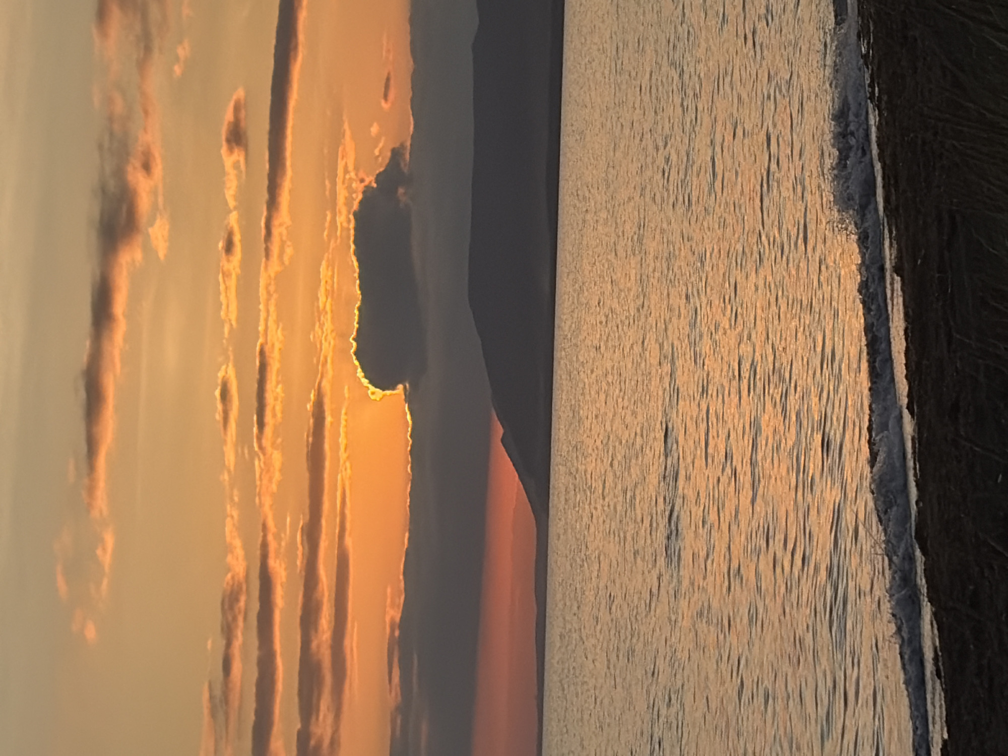 The image depicts a serene sunset over a body of water, likely Lake Ozerskoe, in Ozerskoe, Russia. The sky is ablaze with warm hues of orange and yellow, with scattered clouds adding texture and depth. A dark, silhouetted mountain range stretches across the horizon, partially obscured by a band of reddish-orange light. The water reflects the colors of the sky, creating a shimmering, tranquil surface. A gentle wave laps at the dark, sandy shore in the foreground, with some sparse vegetation visible. There are no people or animals in the image. The scene is one of natural beauty and quietude.
