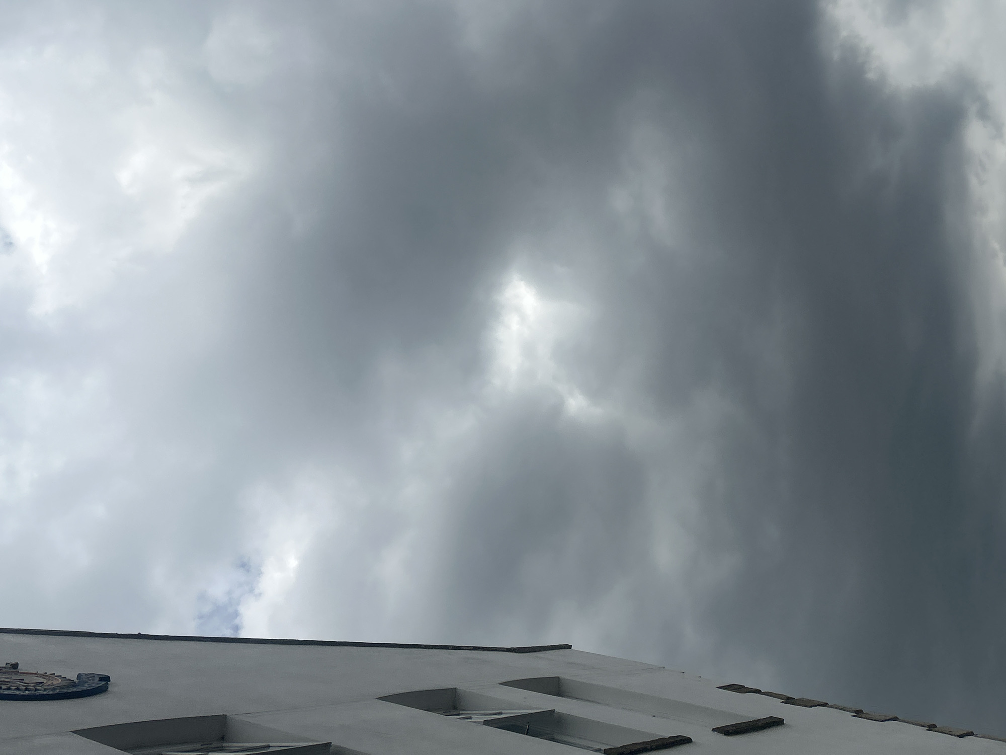This media file captures a close-up, low-angle view of the sky above a white building. The sky is predominantly filled with dark, heavy clouds, suggesting overcast or stormy weather. A small patch of lighter sky, possibly a break in the clouds, is visible towards the center. The building's white facade occupies the lower portion of the image, with a dark, angled roofline separating it from the sky. Notable details on the building include what appears to be a decorative emblem on the left side, and a series of recessed window-like structures or architectural features along the roofline. The lighting is dim and diffused, consistent with a cloudy day. No people are visible. The location context provided is Boston, United Kingdom.