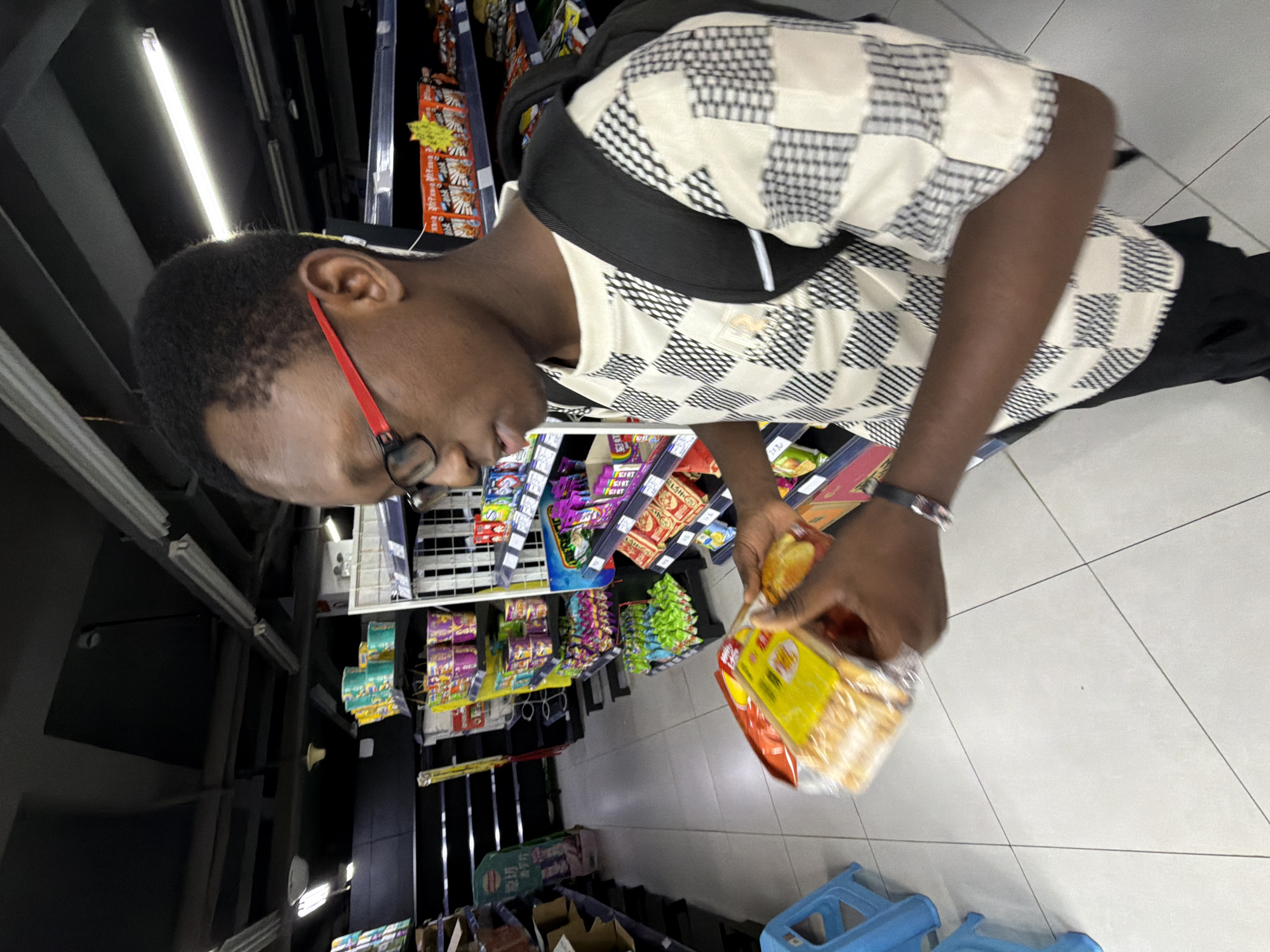 A young Black male wearing glasses and a black backpack stands in a brightly lit grocery store aisle. He is holding a package of what appears to be bread or baked goods in his left hand. He is looking down at the product, examining it. The store shelves are stocked with various packaged food items, including snacks and what look like instant noodles. The flooring is tiled, and the ceiling is equipped with fluorescent lighting. The man is wearing a black and white checkered shirt and black pants. A blue plastic stool is visible to the left of the frame. The scene appears to be a typical retail environment within a city.