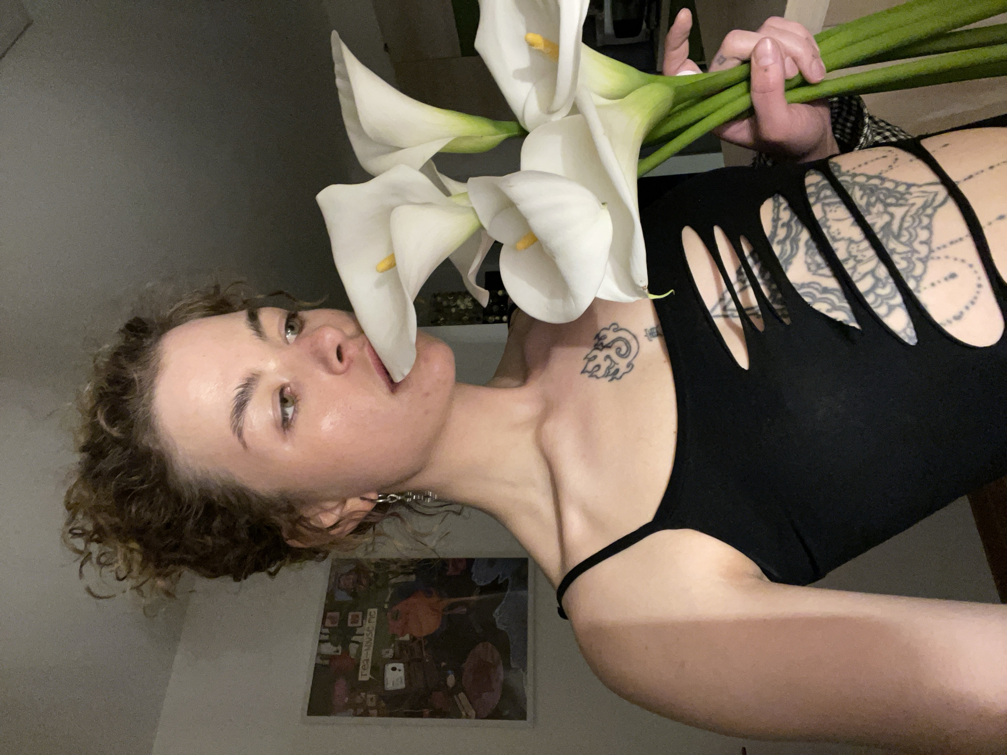 An indoor portrait-style photograph, taken in Boreti, Montenegro, features a person with curly brown hair and green/hazel eyes. The subject is holding a bouquet of several white calla lilies, positioning one of the flowers close to their slightly parted mouth. They are wearing a black top with distinctive cut-outs on the back, which reveal a large, intricate mandala-patterned tattoo on their right shoulder blade. A smaller, abstract tattoo is visible on their left shoulder/collarbone area. The background consists of a plain, light-colored wall. In the lower left corner, a framed picture or collage is visible, on which the partial text "rea-house me" can be discerned. The overall lighting is dim, suggesting an indoor setting under artificial or low natural light, possibly in the evening. The person's expression is neutral and artistic.