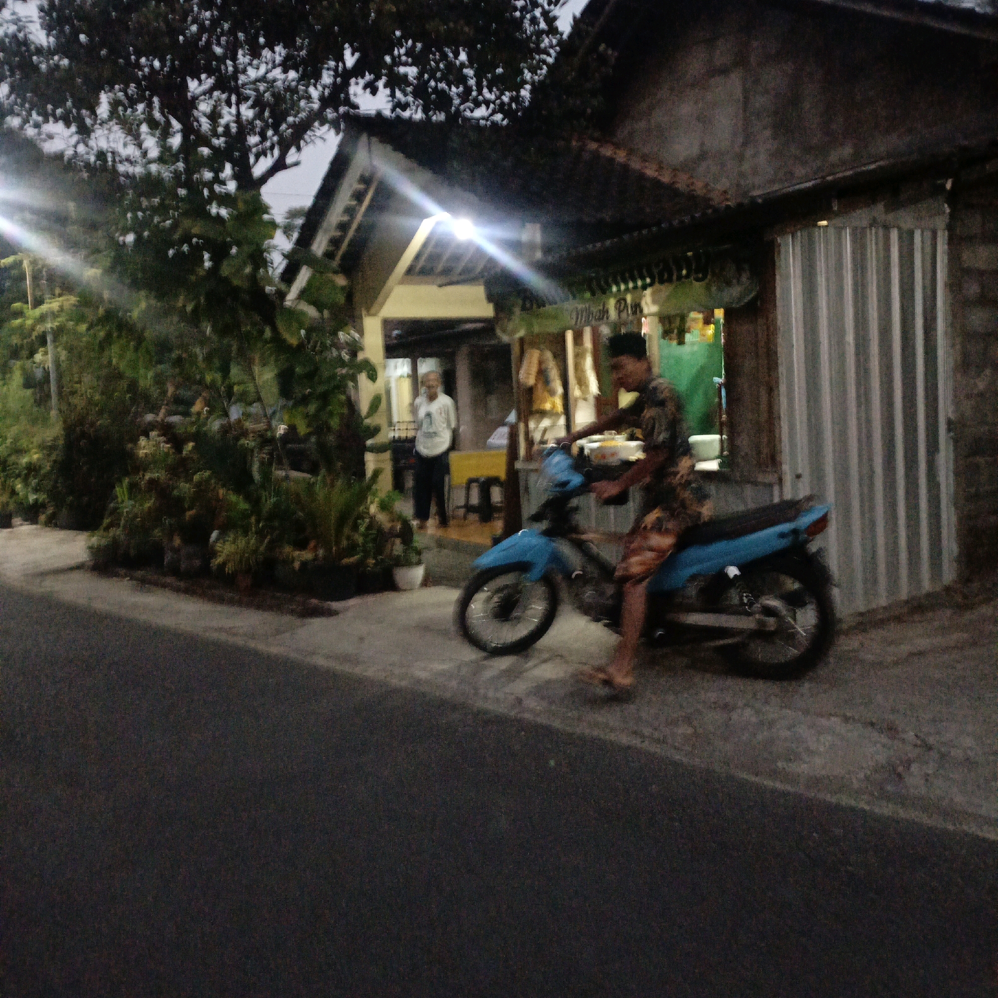 In the city of Salatiga, Indonesia, at dusk, a young man on a blue motorcycle is captured in motion on a paved road in front of a small storefront. The motorcycle is facing right, and the man is wearing a patterned shirt and patterned shorts, with his right foot on the ground for balance. He appears to be about to depart or has just arrived. The storefront behind him features a corrugated metal facade on the right and a more traditional open-fronted shop on the left. Inside the open-fronted shop, an elderly person in a white shirt and dark pants stands near a yellow table and a stool. Lush green plants line the sidewalk in front of the shop, creating a verdant foreground that contrasts with the rustic architecture. The lighting suggests the time is either early morning or late evening, with artificial lights illuminating the storefront.