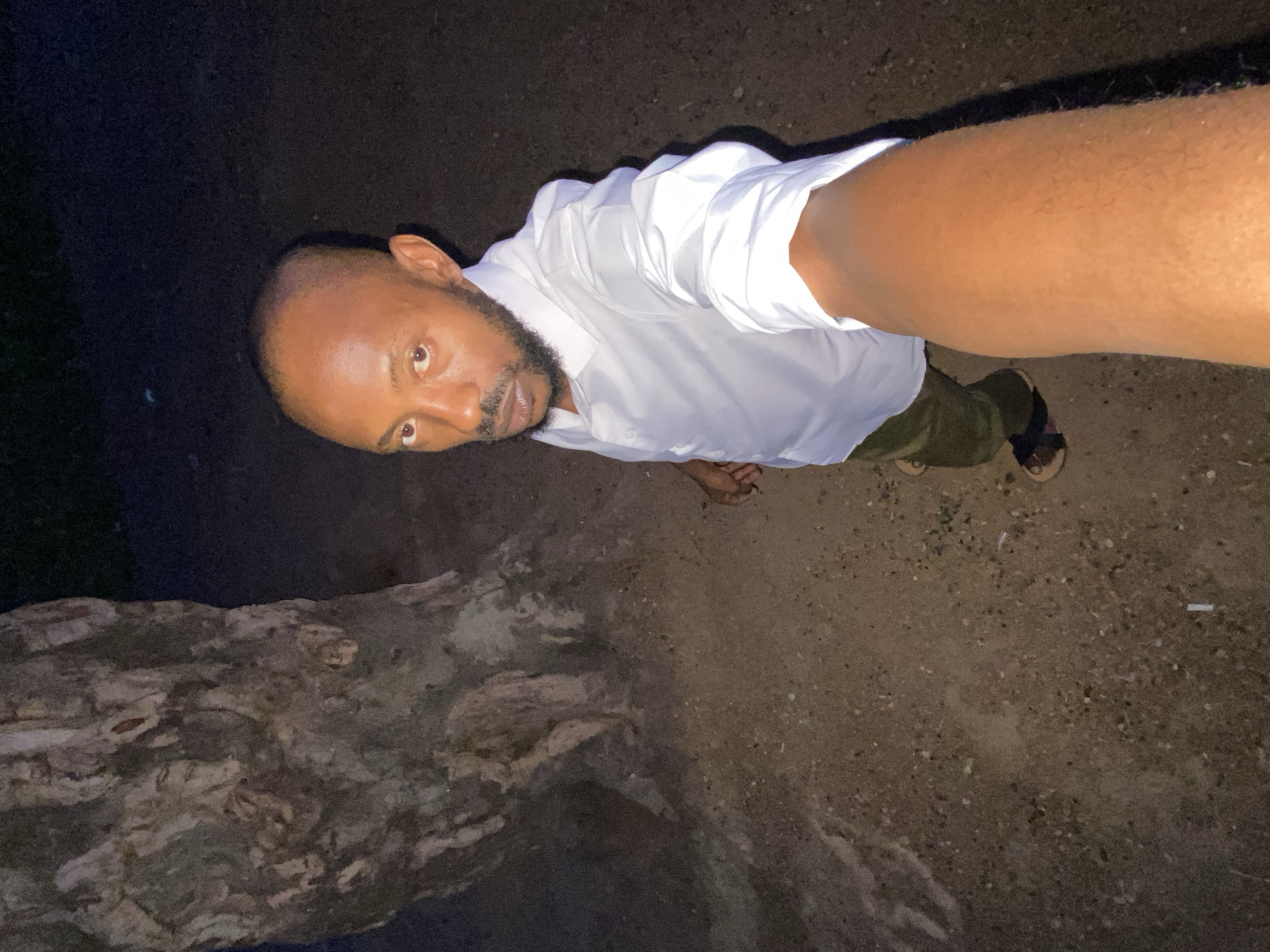 A man with a bald head and a beard is standing on a dirt ground next to a rock formation. He is wearing a white, long-sleeved shirt and olive-green trousers. He also has sandals on. The scene is dimly lit. The location is Sabon Tasha, Nigeria.