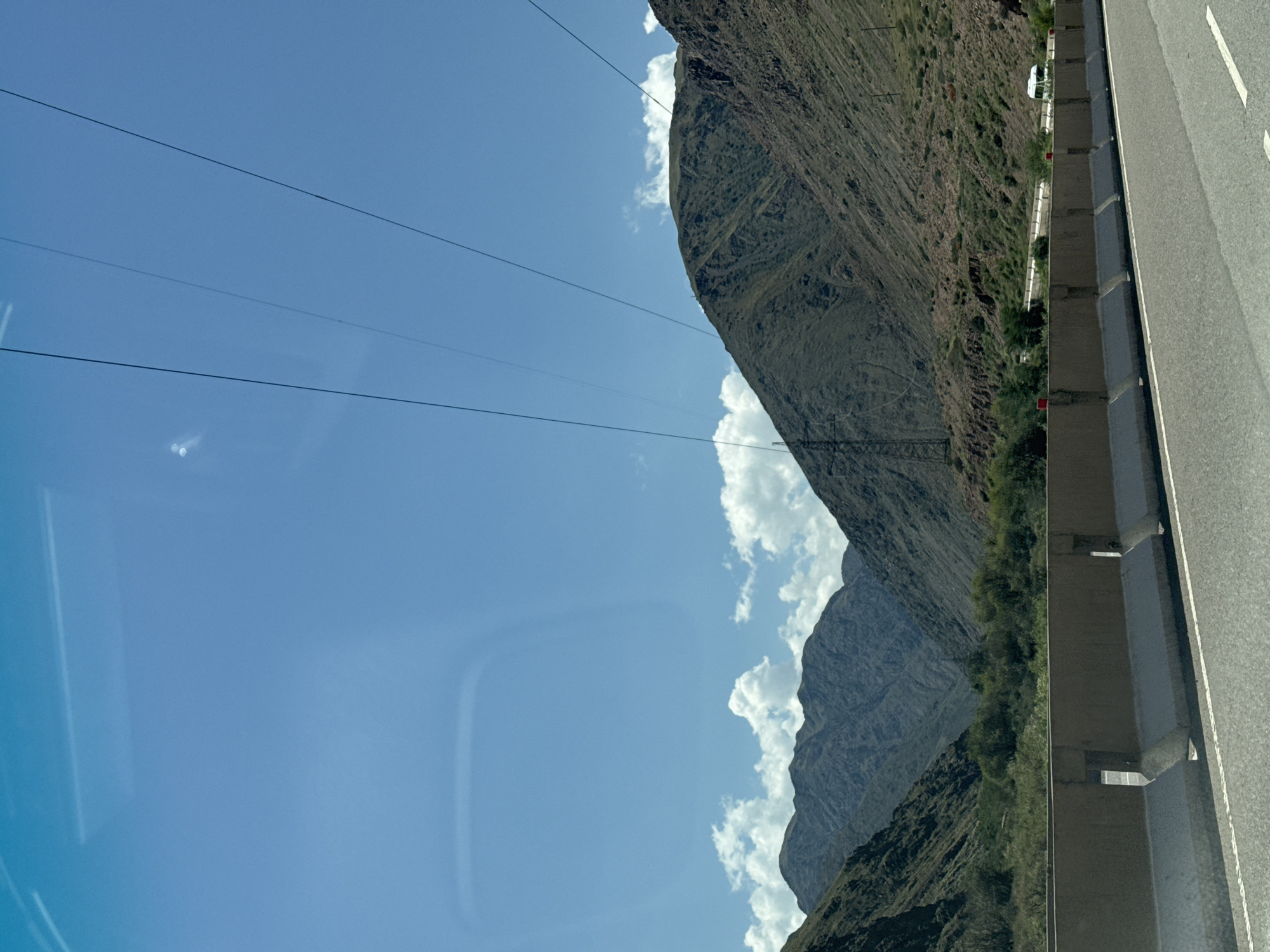 The image shows a mountainous landscape with a clear blue sky and scattered clouds. Several power lines stretch across the sky, passing over the mountain slopes. The mountains are covered in dry vegetation and show signs of erosion. In the foreground, there is a highway with a concrete barrier. A white van is visible on the highway in the distance, driving away from the camera. The scene appears to be a remote, natural area, possibly along a scenic route. The prompt mentions the city of Krasnyy Most, Kyrgyzstan, but this location is not explicitly identifiable from the visual information alone.