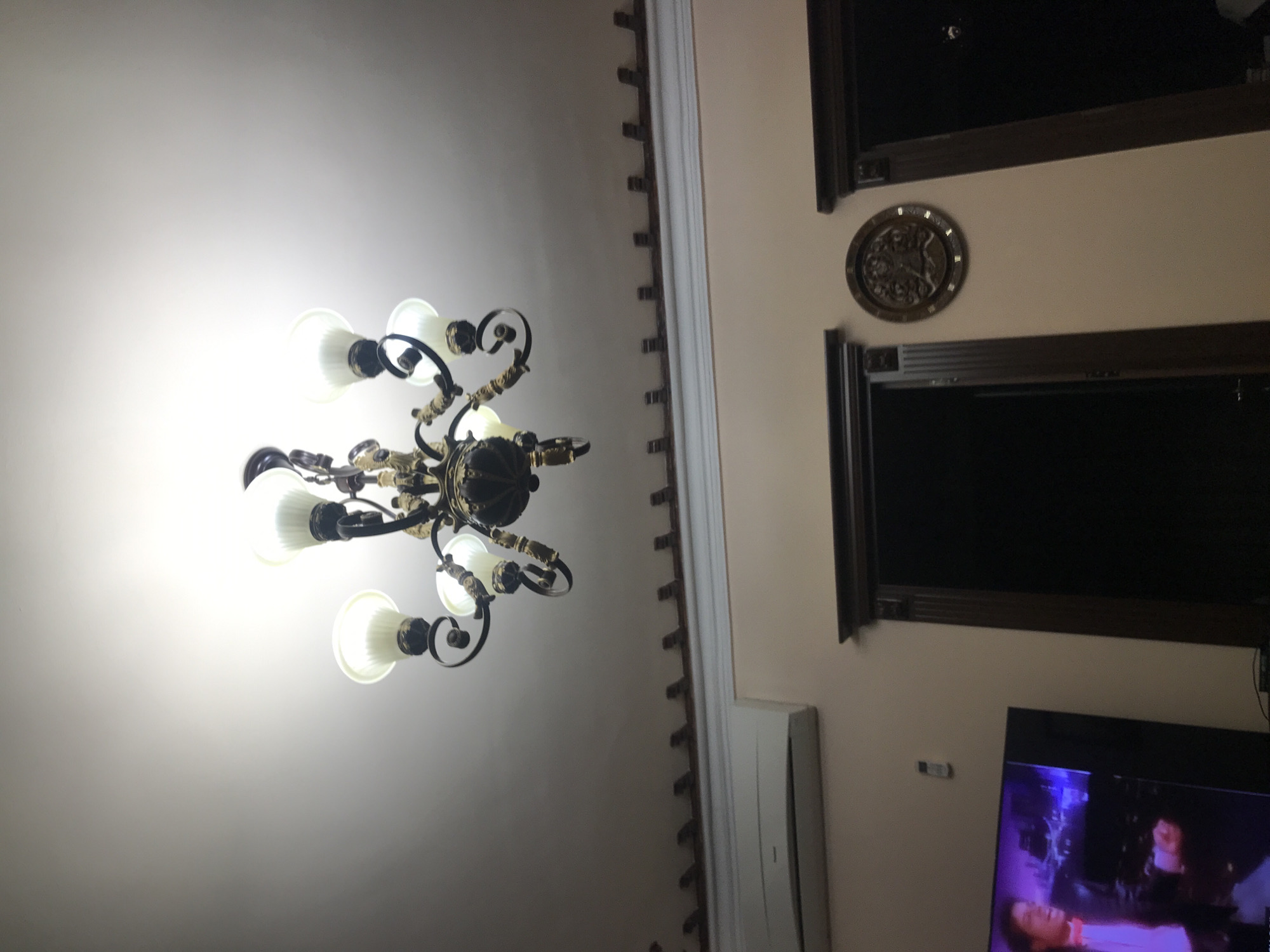 The image shows a room interior. A chandelier is affixed to the ceiling on the left side of the frame. Two framed windows, with closed blinds, are visible on the wall to the right, as well as a decorative medallion. A flat-screen television is situated in the lower right, displaying a scene with people. An air conditioning unit is mounted on the wall to the right of the chandelier. The location is possibly a residential building in Yerevan, Armenia, but that cannot be verified from the image.