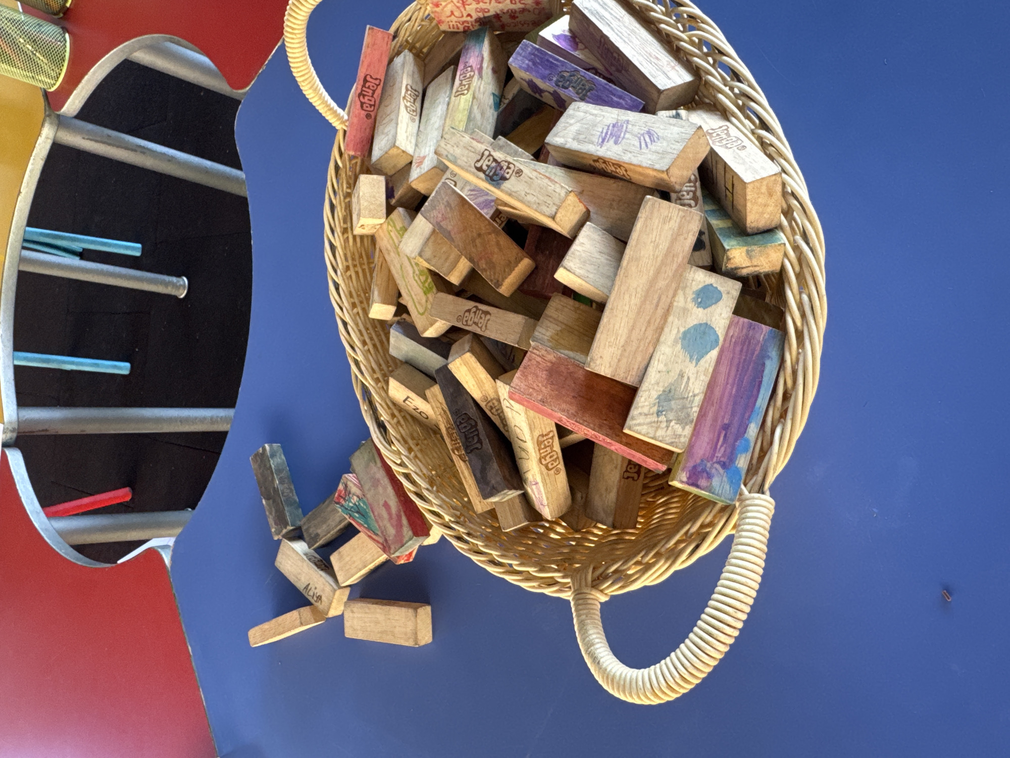 A close-up, top-down shot captures a wicker basket overflowing with wooden Jenga blocks on a vibrant blue table. The Jenga blocks exhibit a variety of colors and designs, with some featuring the "Jenga" logo and others adorned with paint splatters or scribbled marks. A few scattered blocks lie outside the basket on the table's surface. In the background, a curved red tabletop with rounded silver trim is visible, suggesting a playful or recreational setting. The image is devoid of any specific landmarks that would indicate its location in Aksu, Turkey.