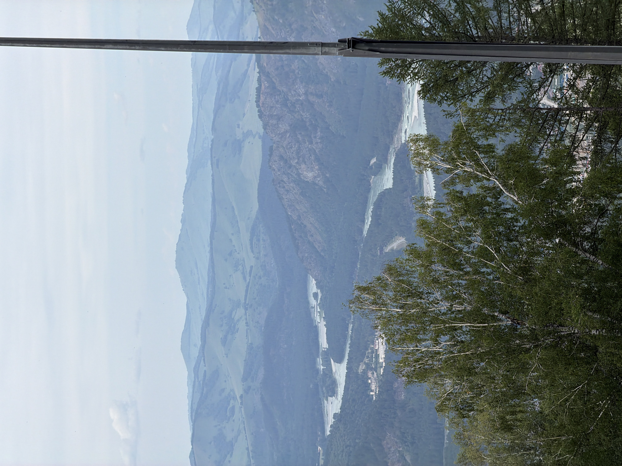 This is a wide, horizontal shot of a vast mountainous landscape with a river winding through it. The foreground is dominated by the lush green foliage of several trees, primarily birch, with their characteristic white bark visible. To the right, a tall, dark gray metal pole, possibly a utility pole or part of a structure, bisects the frame vertically.
The middle ground reveals a wide river, its water a pale turquoise, snaking through a valley densely covered with dark green coniferous trees. On the banks of the river, there are some scattered buildings, appearing as clusters of light-colored structures. Further back, rugged, rocky mountains rise, their slopes also covered in dense forest.
The background consists of rolling hills that gradually fade into a hazy blue-gray distance. The sky is overcast with a pale, uniform light, with a few wispy clouds visible in the upper left quadrant. The overall impression is one of natural grandeur and a remote, tranquil environment. The scene is located in the vicinity of Ozernoe, Russia, likely a mountainous or semi-mountainous region. There are no people or animals visible in the frame.