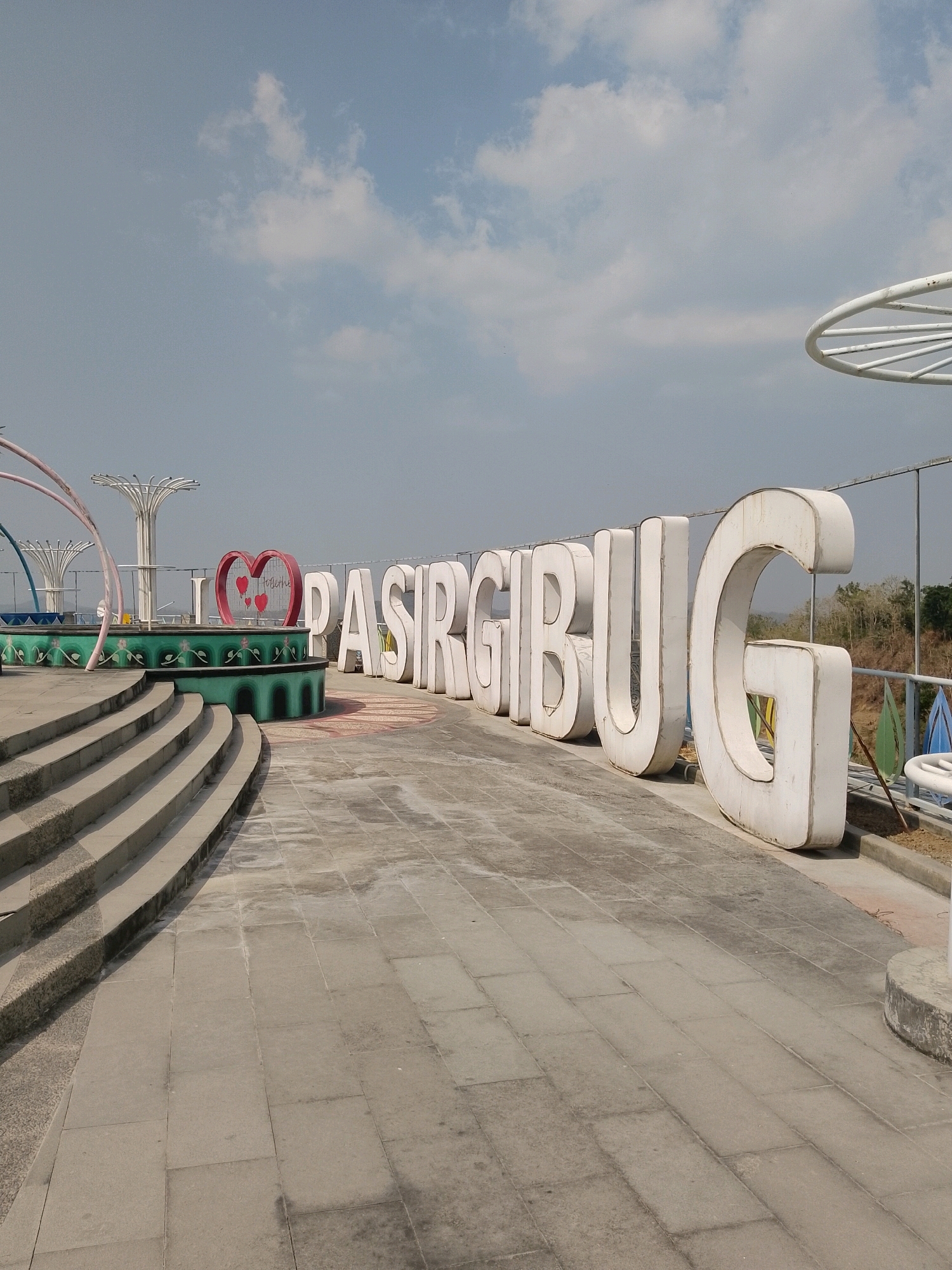 A wide-angle, eye-level outdoor shot captures a scenic view of a public space in Penanggapan, Indonesia. The foreground features a paved walkway with distinct square stone tiles, leading towards a tiered, curved stone staircase on the left. Further back, large, white, three-dimensional letters spell out "I PASIRGIBUG," with a heart symbol between "I" and "PASIRGIBUG." Behind the letters, a decorative green fountain structure is visible, adorned with intricate patterns. In the background, a metal railing separates the walkway from a gentle slope covered in sparse vegetation. The sky above is partly cloudy, with scattered white clouds against a pale blue backdrop. The overall environment suggests an outdoor recreational area or tourist attraction. There are no people visible in the frame.