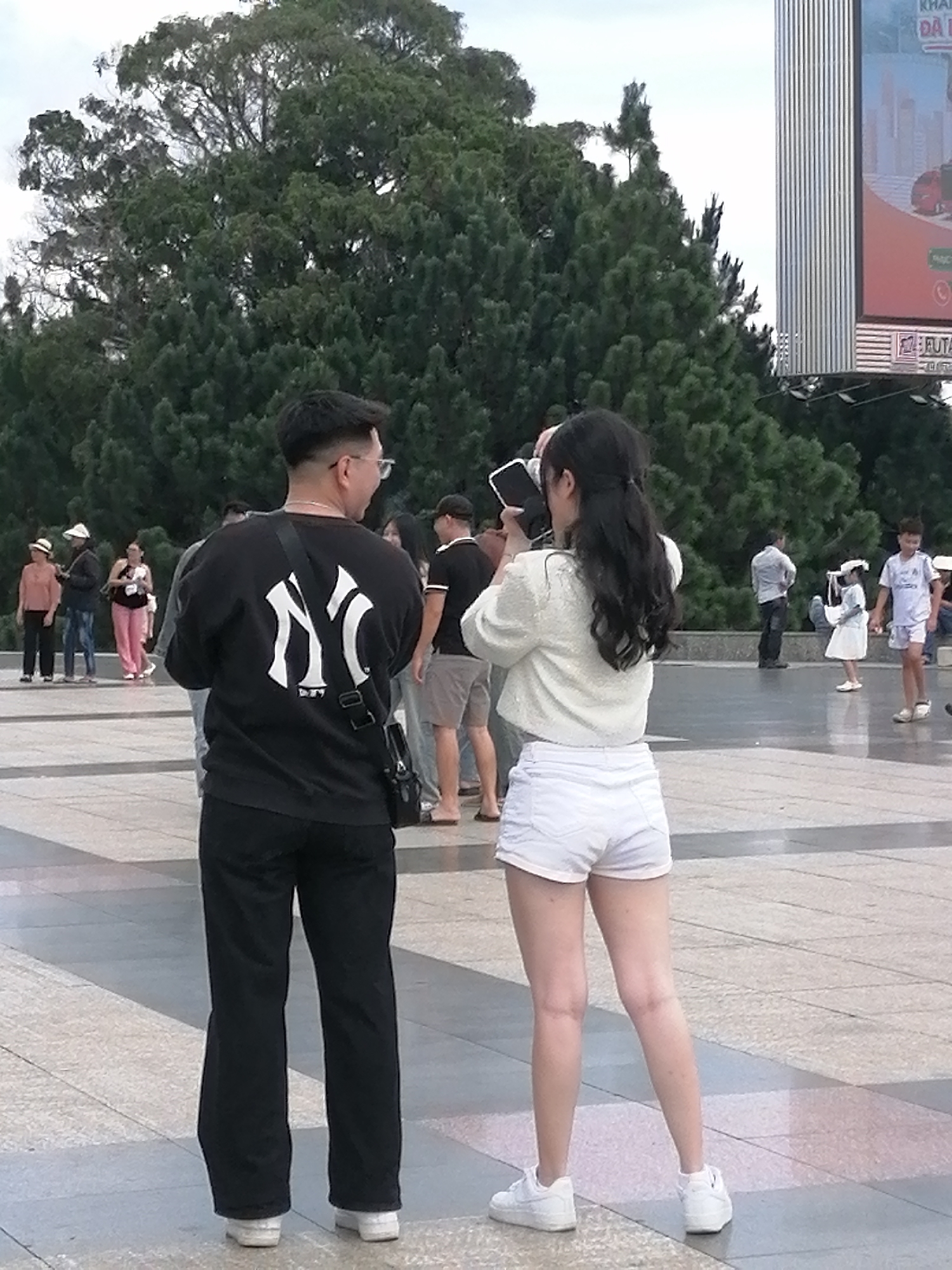 The image depicts an outdoor urban scene, likely in Da Lat, Vietnam, given the presence of a prominent billboard with Vietnamese text. In the foreground, a man wearing a black sweatshirt with a white "NY" logo and black pants stands with his back to the camera, facing a woman. The woman, also with her back to the camera, is wearing a light-colored, textured top and white shorts. She is holding up a smartphone, appearing to take a photograph. Behind them, a large tree with dense green foliage dominates the background, with a tall, ribbed advertising billboard to the right. Several other individuals are visible further in the background, including a person in a black t-shirt and shorts, a child in a white dress and hat, and another child in athletic wear. The ground is paved with large, light-colored square tiles. The overall atmosphere suggests a public gathering or tourist area.