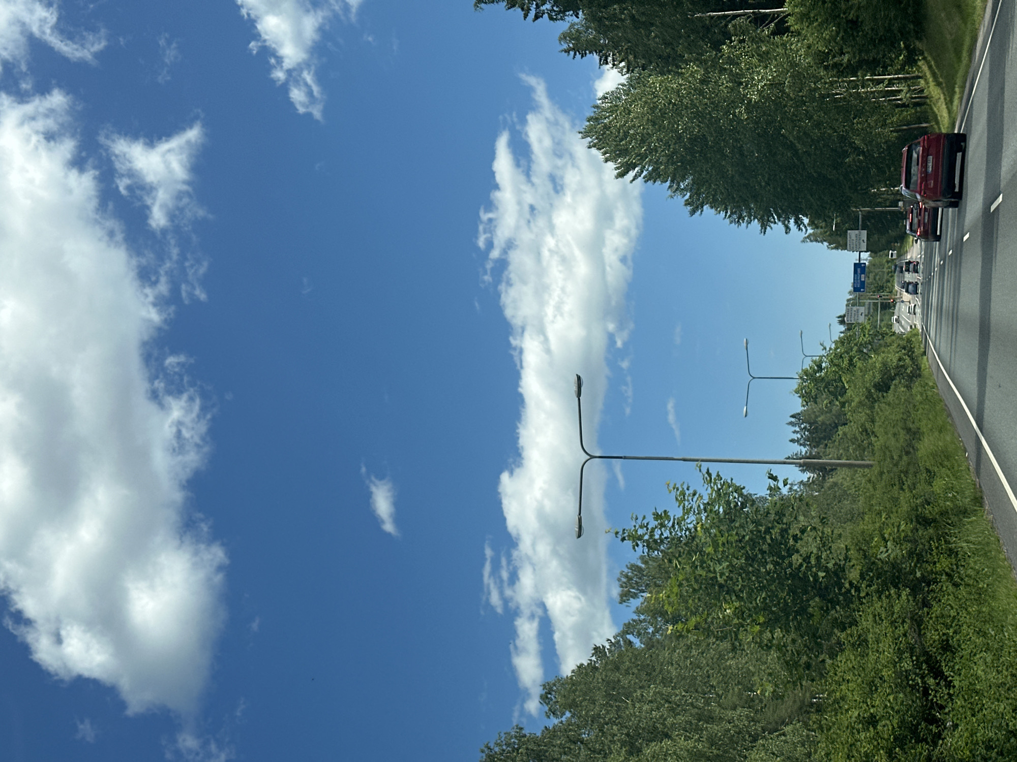 The image depicts a roadside view on a sunny day, with a clear blue sky dotted with white clouds. The perspective suggests the photo was taken from a moving vehicle, looking forward and slightly to the left. Dense green trees line both sides of a multi-lane highway, which stretches into the distance. Several cars are visible on the road, including a red vehicle in the foreground. Streetlights stand at intervals along the highway. There are road signs with text, but it is illegible in the image. The overall impression is one of a typical drive through a verdant, possibly rural or suburban, landscape.