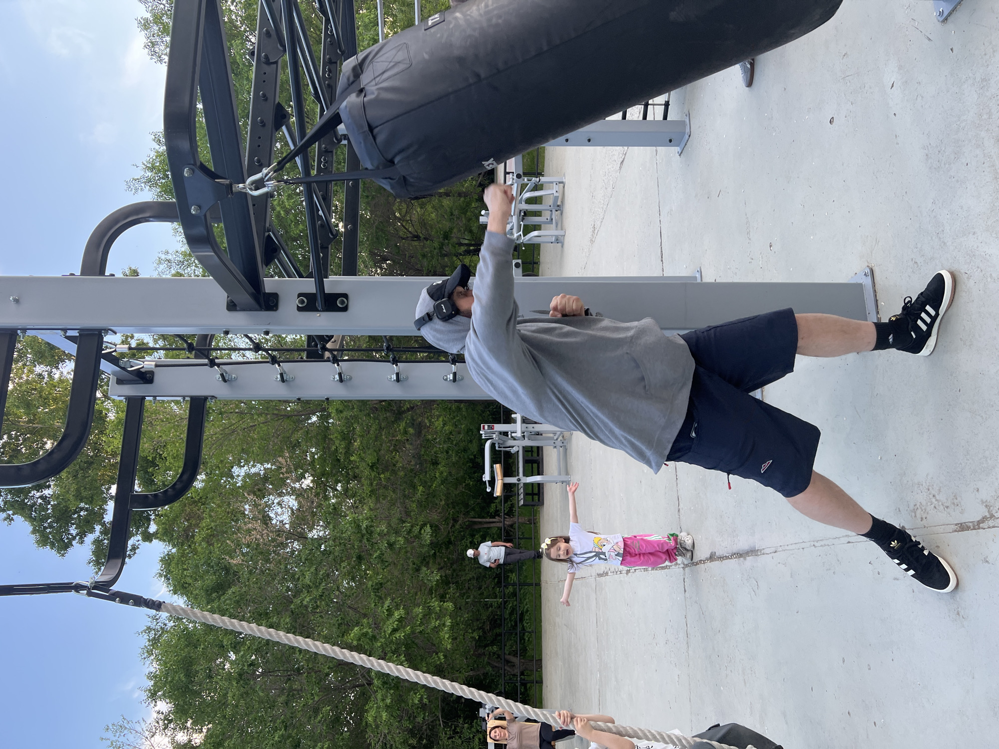 In Khabarovsk, Russia, an outdoor fitness park features a man in a grey hoodie and dark shorts wearing headphones and black sneakers with white stripes. He is actively punching a large black punching bag hanging from a metal frame. Nearby, a young girl in a white t-shirt with cartoon characters and pink pants stands with her arms outstretched. In the background, other individuals are visible, some engaging with exercise equipment, and a person stands near trees. The ground is paved, and the environment suggests a public recreational area with a backdrop of lush green trees under a bright sky.