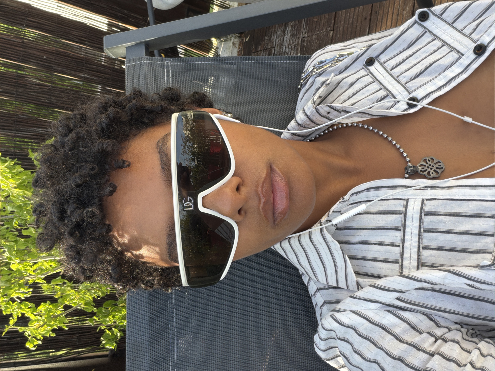 The image shows a person relaxing outdoors in Champs-sur-Marne, France. The subject, a young adult with dark, tightly curled hair and a medium-dark skin tone, is seen from a slightly elevated, close-up perspective, lying back on a dark gray mesh outdoor chair.
They are wearing a light-colored, possibly linen, button-down shirt with thin black vertical stripes, which is unbuttoned. White-framed sunglasses with dark lenses cover their eyes; the letters "DG," likely a brand logo, are visible on the left lens. A silver beaded necklace with an ornate, possibly floral or geometric, pendant rests on their chest. White wired earphones are draped around their neck and upper chest. Their lips are slightly parted, revealing a subtle gloss.
The background features vibrant green foliage, suggesting bushes or trees, and a dark, horizontal slatted fence or screen, characteristic of a garden, patio, or balcony setting. Bright sunlight illuminates the scene, casting reflections on the sunglasses and creating clear highlights on the person's skin and accessories. The overall impression is one of leisure and calm during what appears to be a sunny day.