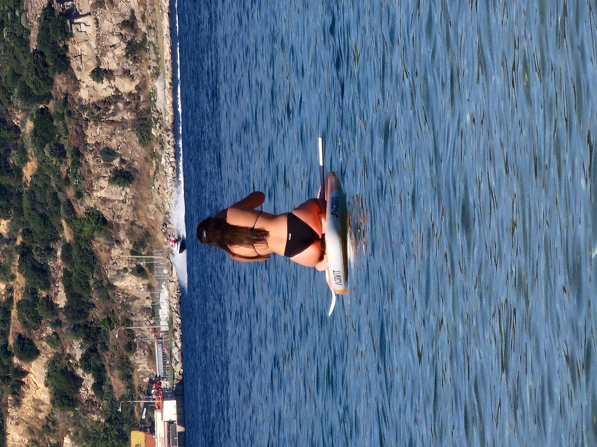A woman in a black bikini is kneeling on a paddleboard in the blue ocean. Her back is to the camera, and her long, dark hair is cascading down her back. The paddleboard has an orange and white design and the word "ACASA" printed on its side. In the background, a rocky cliff covered in green vegetation rises from the water's edge. A jet ski is speeding across the water, leaving a white wake behind it. A dock with shipping containers and lights is visible on the shore. The scene appears to be set in Nea Peramos, Greece, with the distinctive Mediterranean landscape.