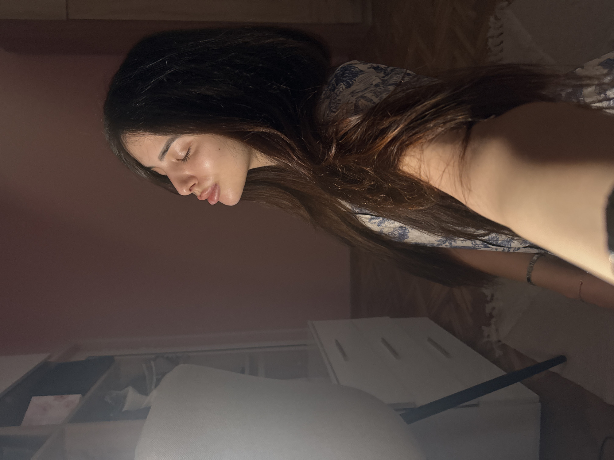 The image shows a young woman with long, dark hair, wearing a blue and white patterned top. She is positioned in a profile view, with her eyes closed and her lips slightly parted, appearing to be in a moment of reflection or relaxation. She has a bracelet on her left wrist. The background suggests an indoor setting, possibly a bedroom or living area, with a dresser or wardrobe visible on the left and a pink-colored wall behind her. The lighting is warm and somewhat dim, casting shadows and highlighting her face. The floor appears to be wooden with a rug. There is no indication of the city of Plovdiv, Bulgaria.