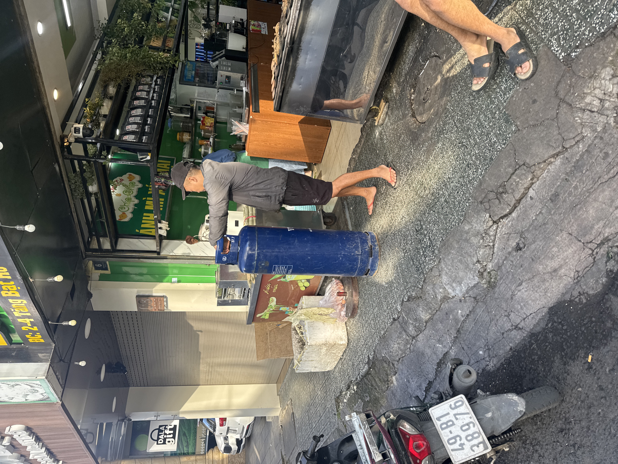 A man wearing a baseball cap, a grey jacket, black shorts, and flip-flops is holding a large blue propane tank with a handle. The tank is standing upright. The scene appears to be outdoors on a street in Da Lat, Vietnam. In the background, there is a storefront with a green awning and shutters. There are also motorcycles parked on the street, and some people's legs are visible in the foreground, indicating they are sitting down. The ground is paved with cracked concrete and textured tiles.
