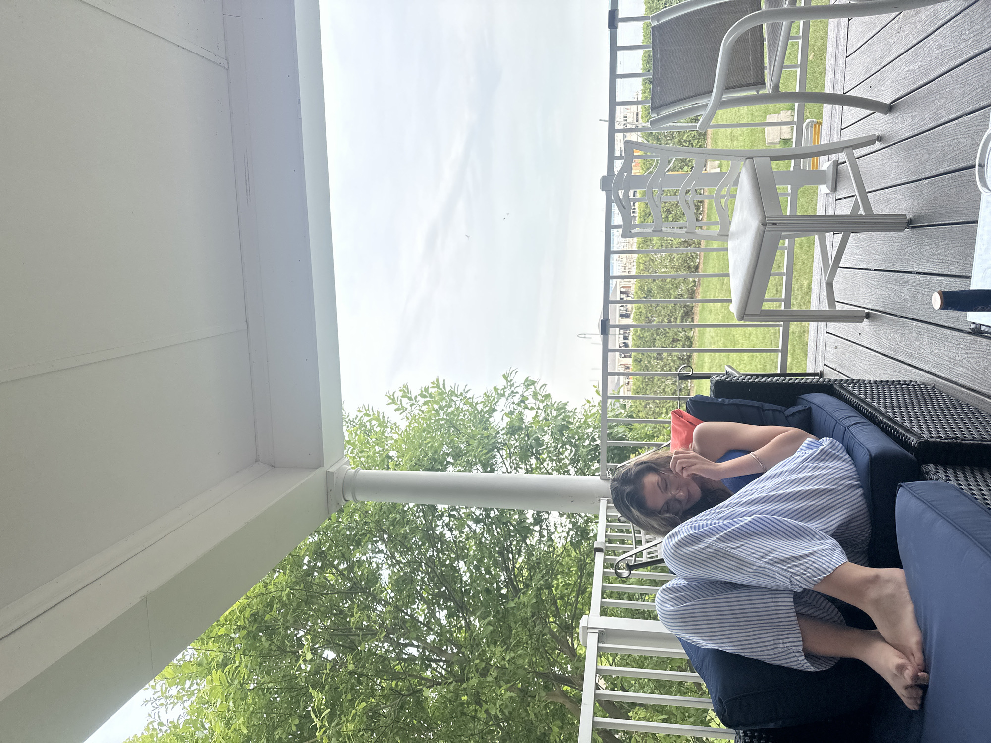 A young woman with long brown hair and glasses, wearing a blue tank top and striped pajama pants, sits cross-legged on a dark blue outdoor couch on a porch. She is looking down and touching her face. The porch has a white railing and a white roof supported by a white pillar. Beyond the railing, there is a green lawn, more white railings, and some buildings in the distance under a cloudy sky. The porch floor is made of grey composite decking. There are two white chairs and a grey lounge chair on the deck.