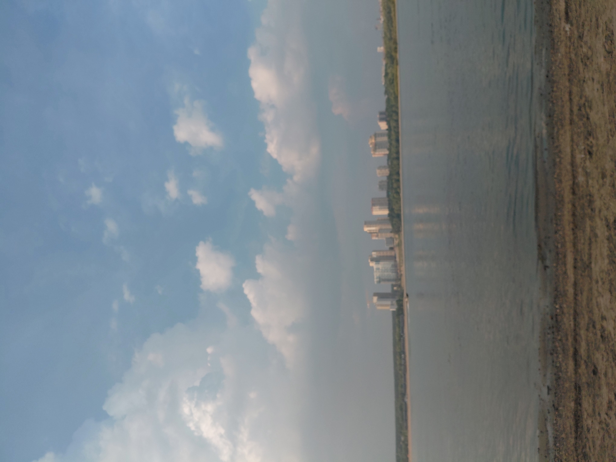 This image depicts a wide shot of the Ob River in Novosibirsk, Russia, taken from a sandy shore in the foreground. In the middle ground, the river stretches across the frame, with a distant embankment lined with lush green trees. Beyond the trees, a cluster of modern, high-rise buildings, indicative of a city skyline, is visible. The sky above is a pale blue, dotted with fluffy white clouds, suggesting a clear, pleasant day. A small boat is present on the water, moving towards the left side of the frame. The overall scene conveys a sense of urban development meeting natural landscape.