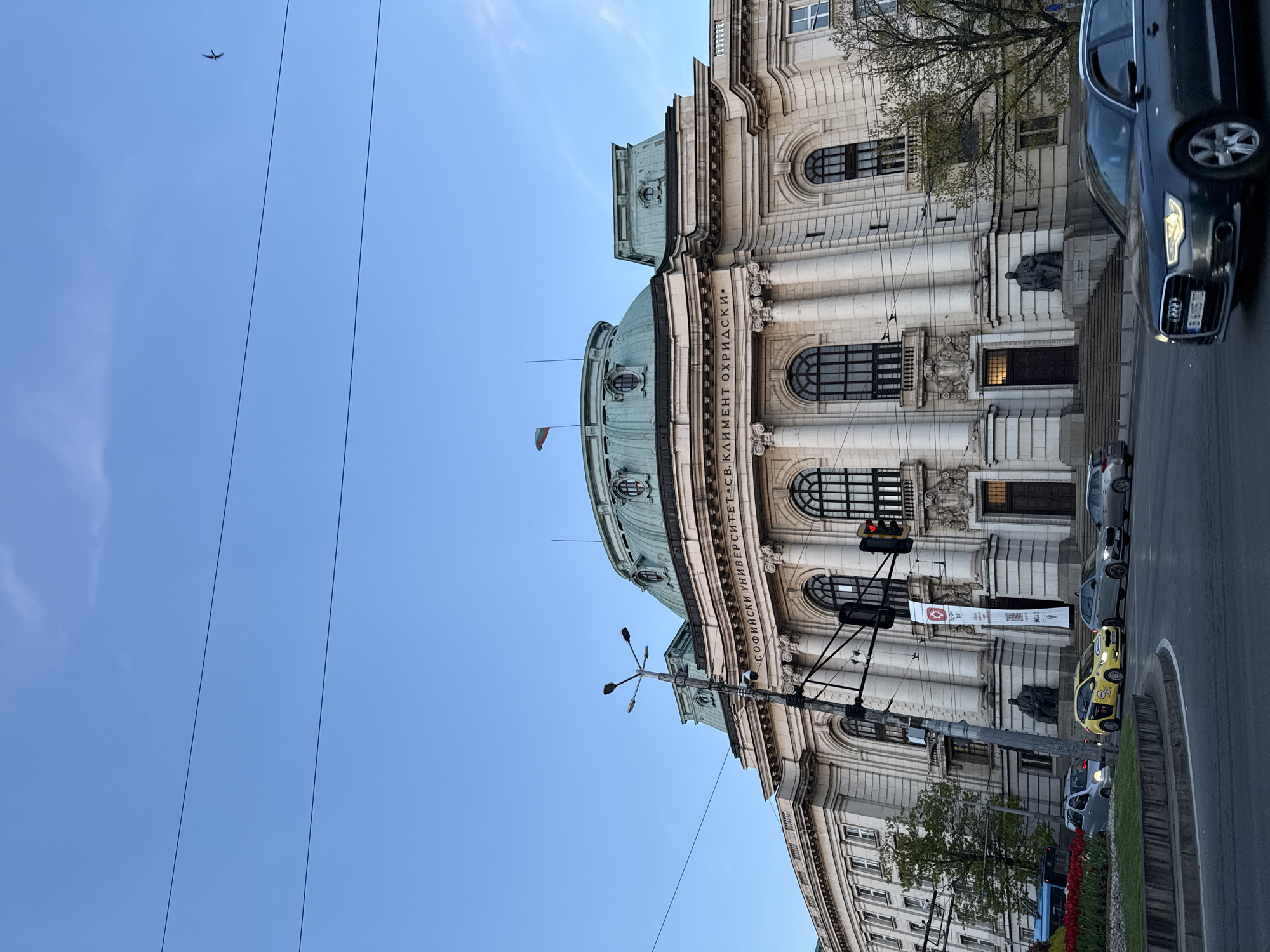 The image presents a daytime street scene in Sofia, Bulgaria, dominated by the grand, classical architecture of Sofia University "St. Kliment Ohridski." The majestic building, constructed from light-colored stone, features prominent columns, arched windows, and a distinctive green copper dome. The inscription "СОФИЙСКИ УНИВЕРСИТЕТ "СВ. КЛИМЕНТ ОХРИДСКИ"" is clearly visible along its facade. A dark statue, likely depicting historical figures, stands at the base of the university's entrance steps.
In the foreground, a multi-lane street carries traffic, with several vehicles visible. A dark grey Audi sedan is prominent on the right, partially cropped, while a yellow taxi and other parked cars can be seen further down the road. Overhead power lines or trolley lines stretch across the frame.
The sky is a clear, bright blue, indicating good weather, and a small bird is captured flying high above. The lighting suggests it is daytime, possibly late afternoon. Street furniture such as a traffic light (displaying red), a multi-armed lamppost, and a pole with a white banner are present. The banner includes the text "ESG UP 3.0" and a QR code. Green trees with developing foliage and flower beds filled with red and yellow blooms add touches of nature to the urban environment. The scene depicts a typical, vibrant day in a historic city center.